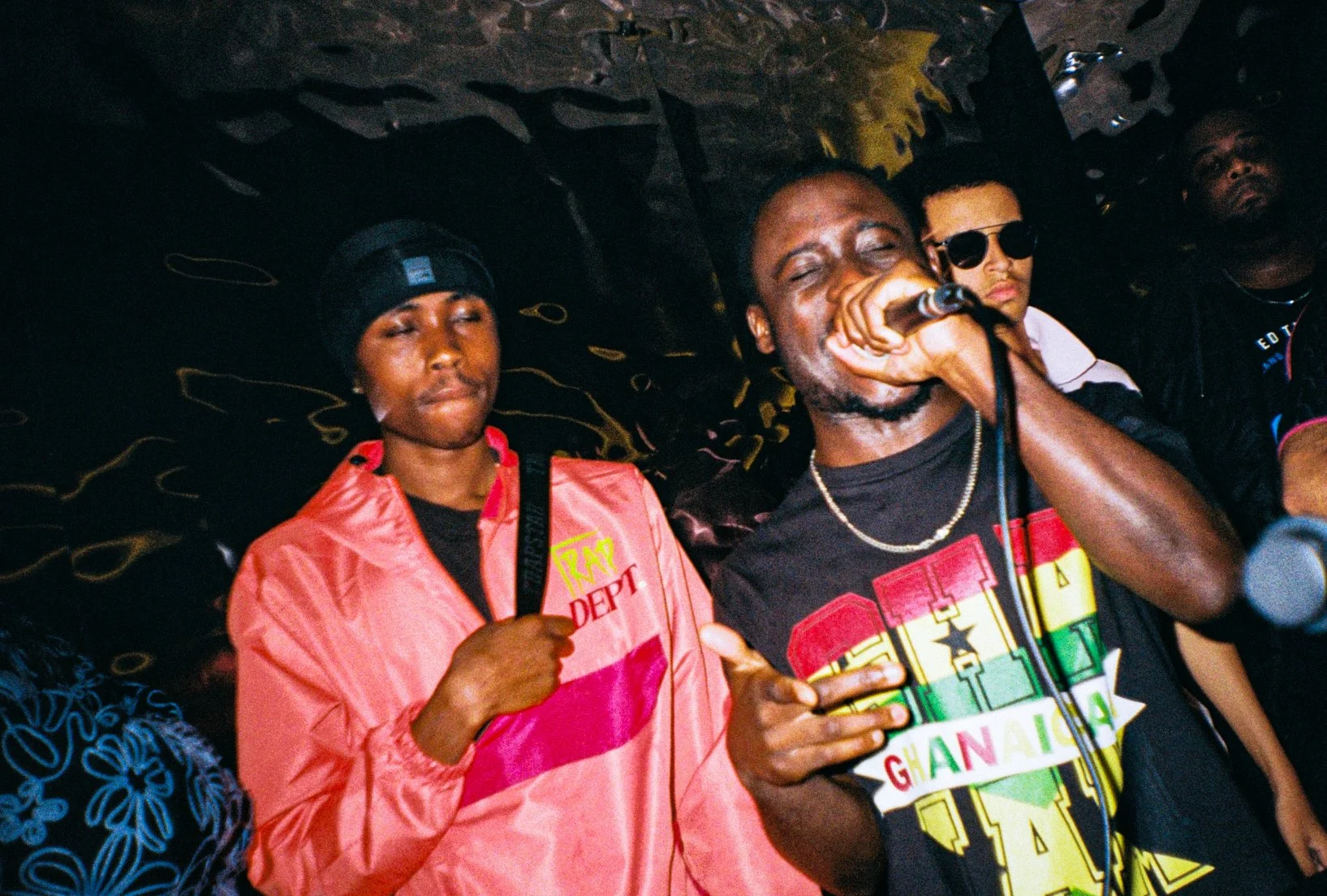 A group of four people at a music event. The person in the front right is singing into a microphone, wearing a colorful Ghana-themed T-shirt. Next to him, a woman with a black cap and pink jacket is holding a strap, with her eyes closed. Two other in