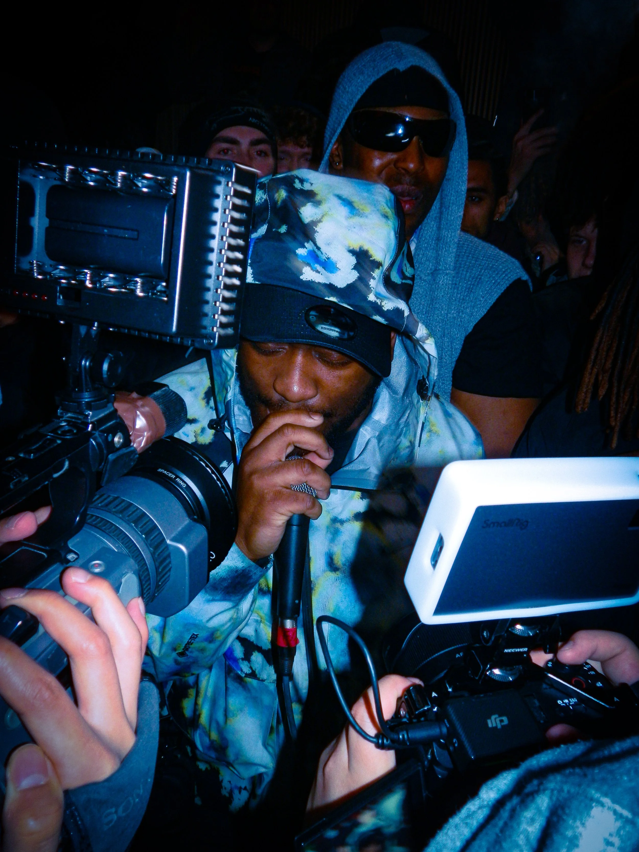 Mc Renz performing holding a microphone with cameras and a crowd surrounding him at a travs presents rave 