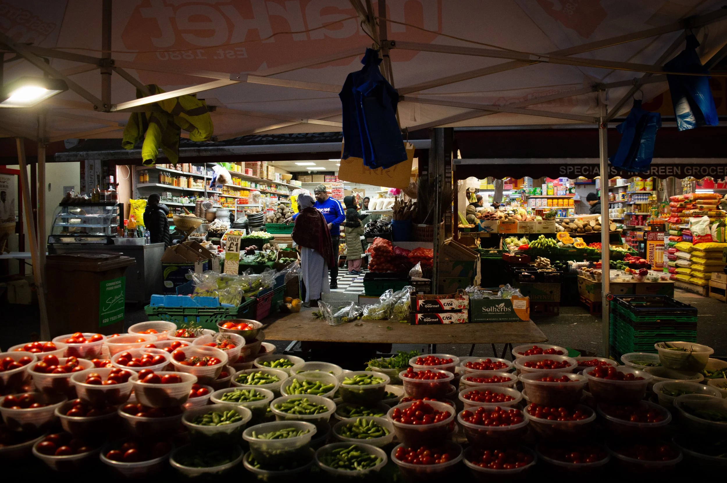low lighting landscape of a fresh produce market stall at East Street Market with a view into the ethnic grocery shop behind it at night