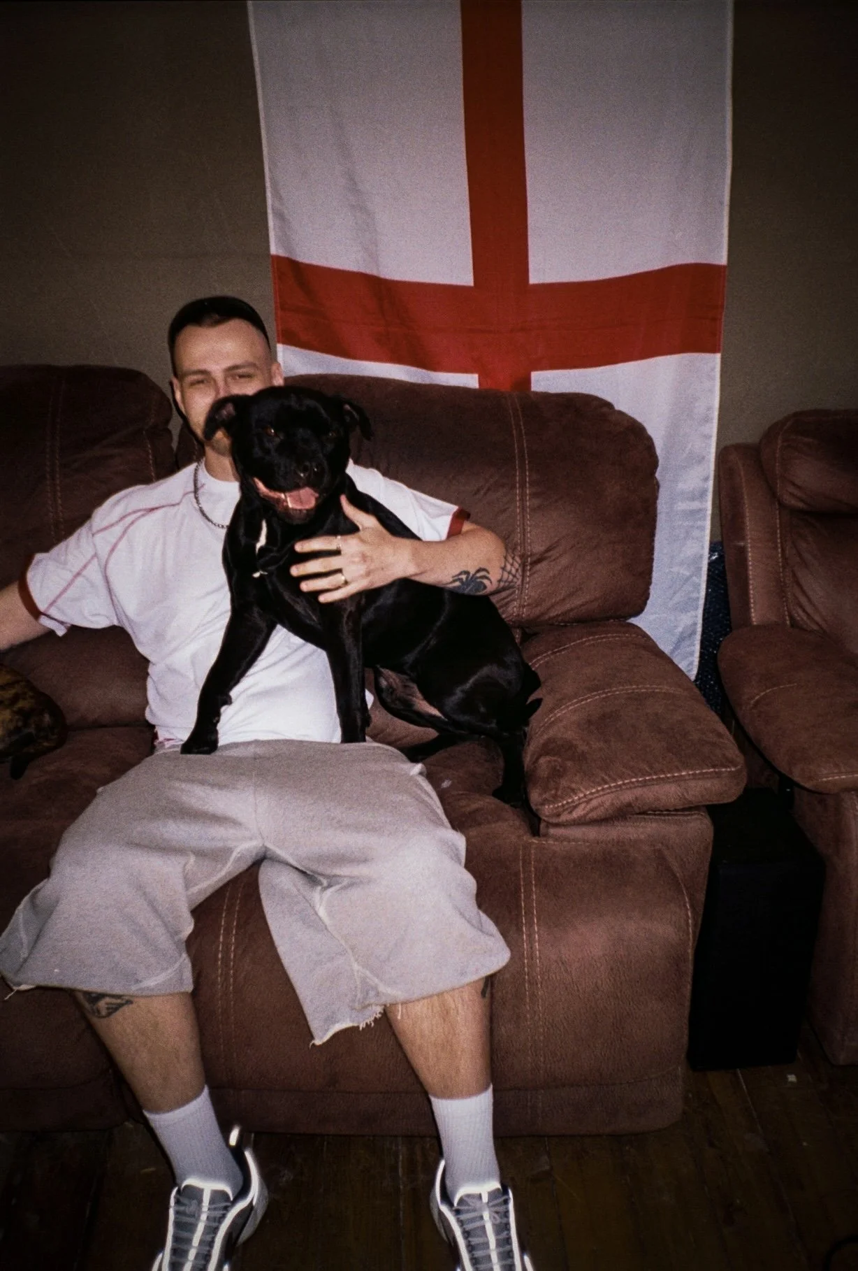 male fashion model wearing defender sportswear sat on a sofa with aa staffy dog and the England flag behind him