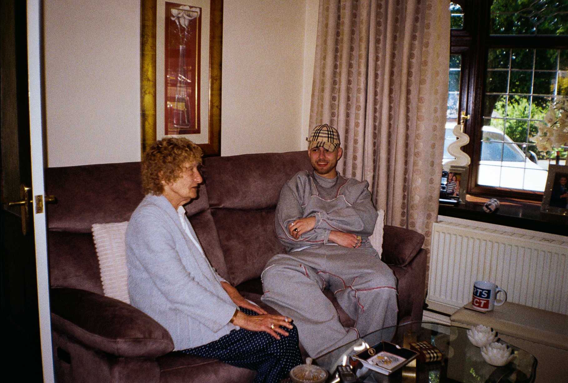 fashion campaign image of young man sat with an elderly woman in her house, wearing defender sportswear garments and Burberry cap