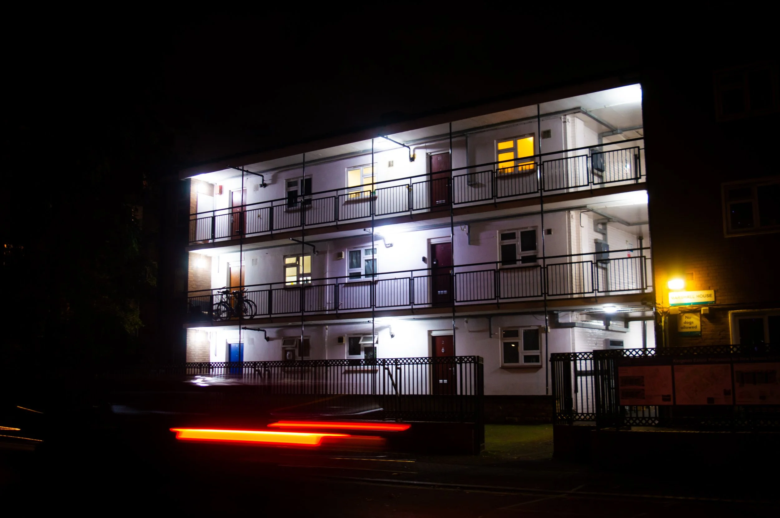 council estate with bright lights above the doors at night and a car driving past