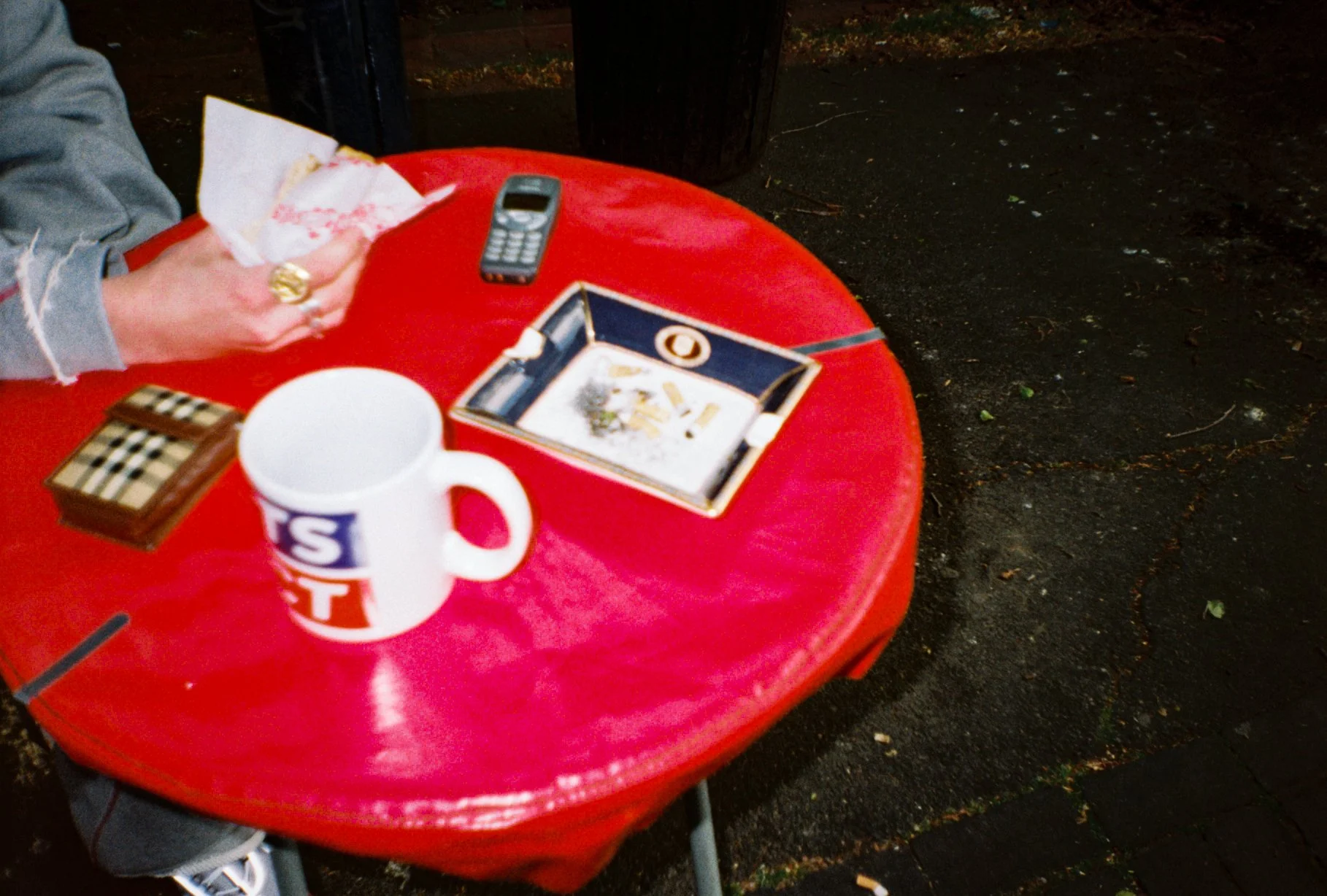 landscape detail photo of a burger van red table, on the table is a sports direct mug, a Burberry ashtray, Burberry cigarette case, Nokia phone and a mans hand holding a burger