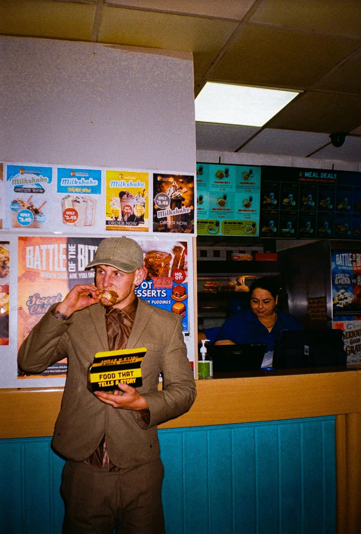 young man wearing beige/brown toned suit and a baseball style cap eating chicken and chips at a chicken shop counter