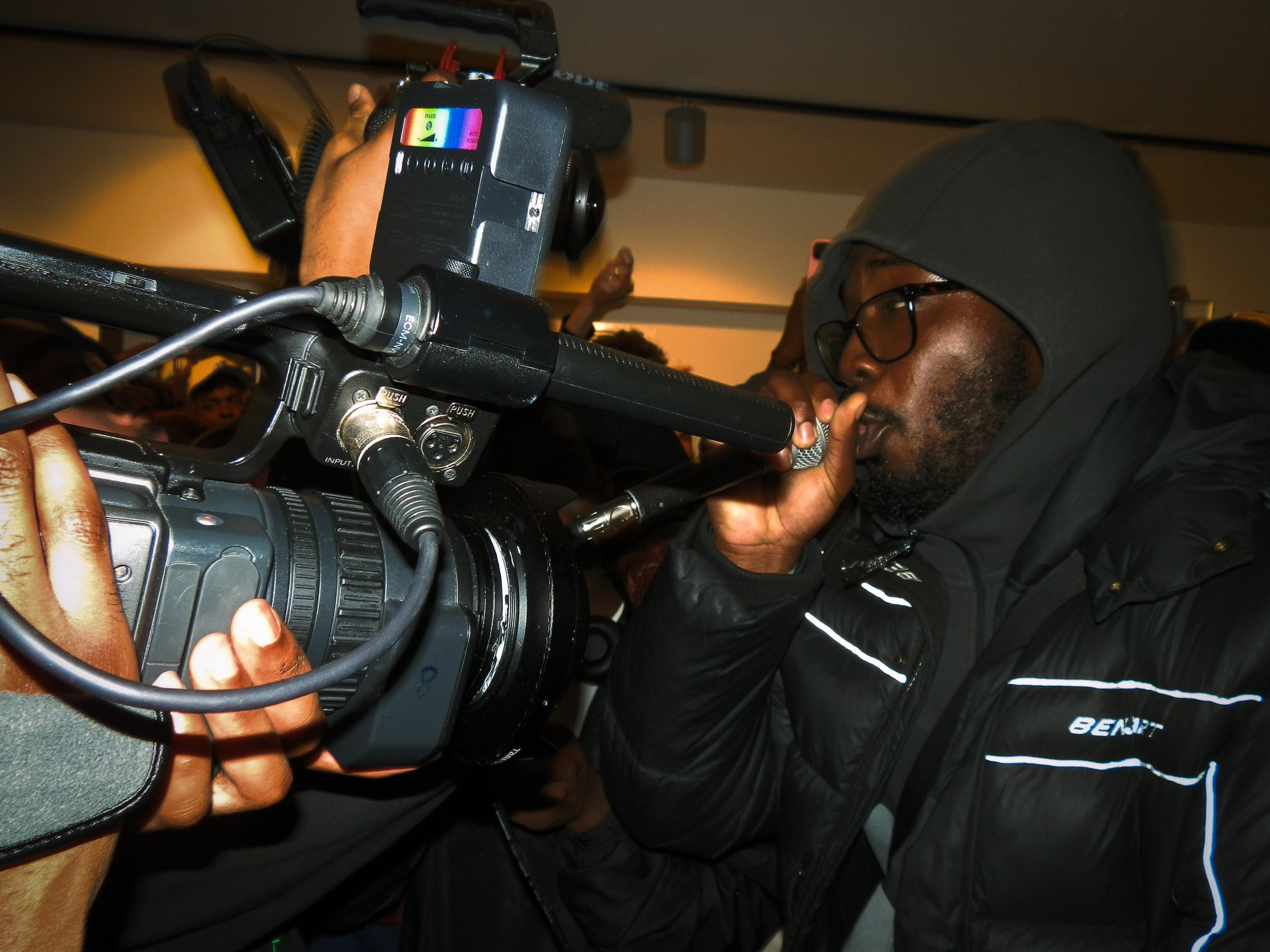 an Mc performing into a cam corder with a mic in hand at the Simon Wheatley pop up with victory lap 