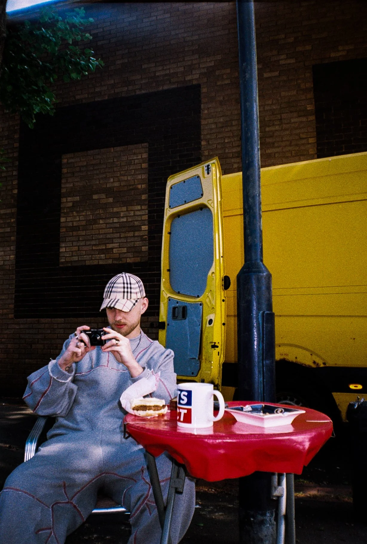 Male fashion model looking at a digital camera sat at a burger van table, yellow van in the background