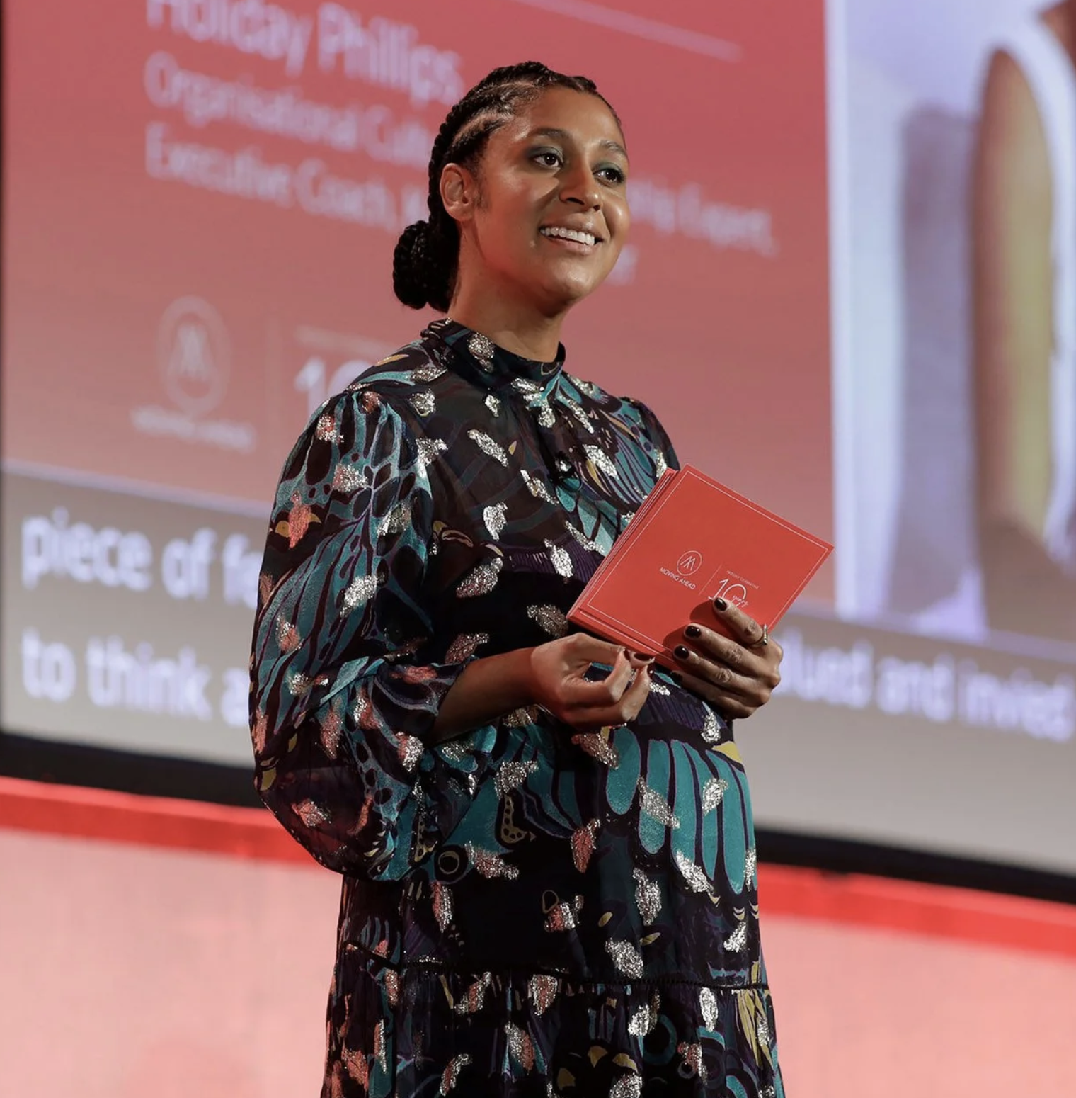 A woman with braided hair wearing a colorful patterned dress, standing on stage, holding a red booklet, smiling during a presentation.