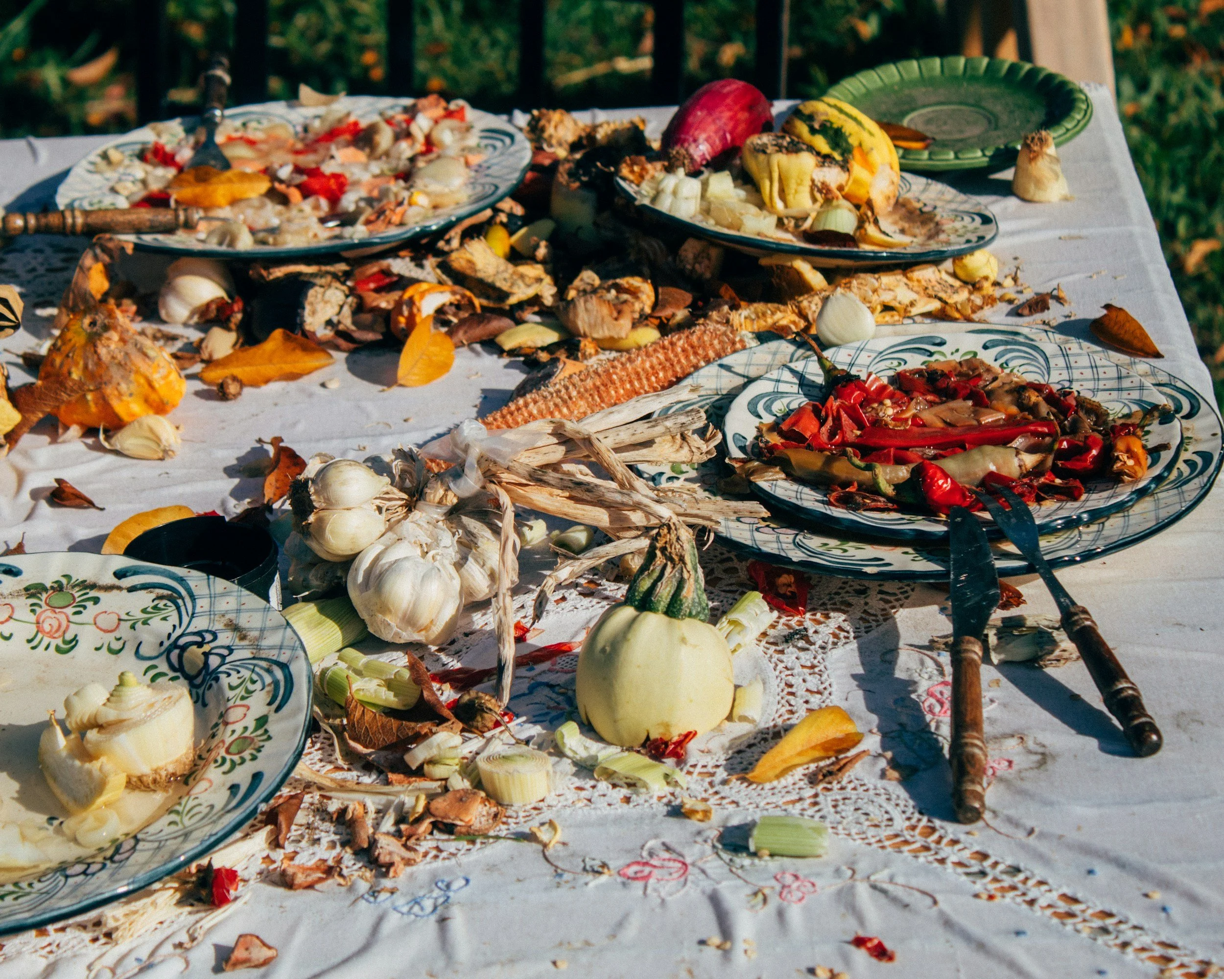 A table outdoors with various leftover plates of food, garlic bulbs, a small decorative gourd, and scattered autumn leaves after a meal, possibly during fall.