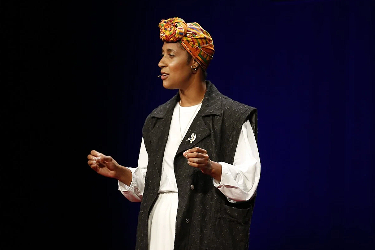 A woman wearing a colorful headwrap, a white dress, and a dark vest, standing on a dark stage and speaking into a headset microphone.