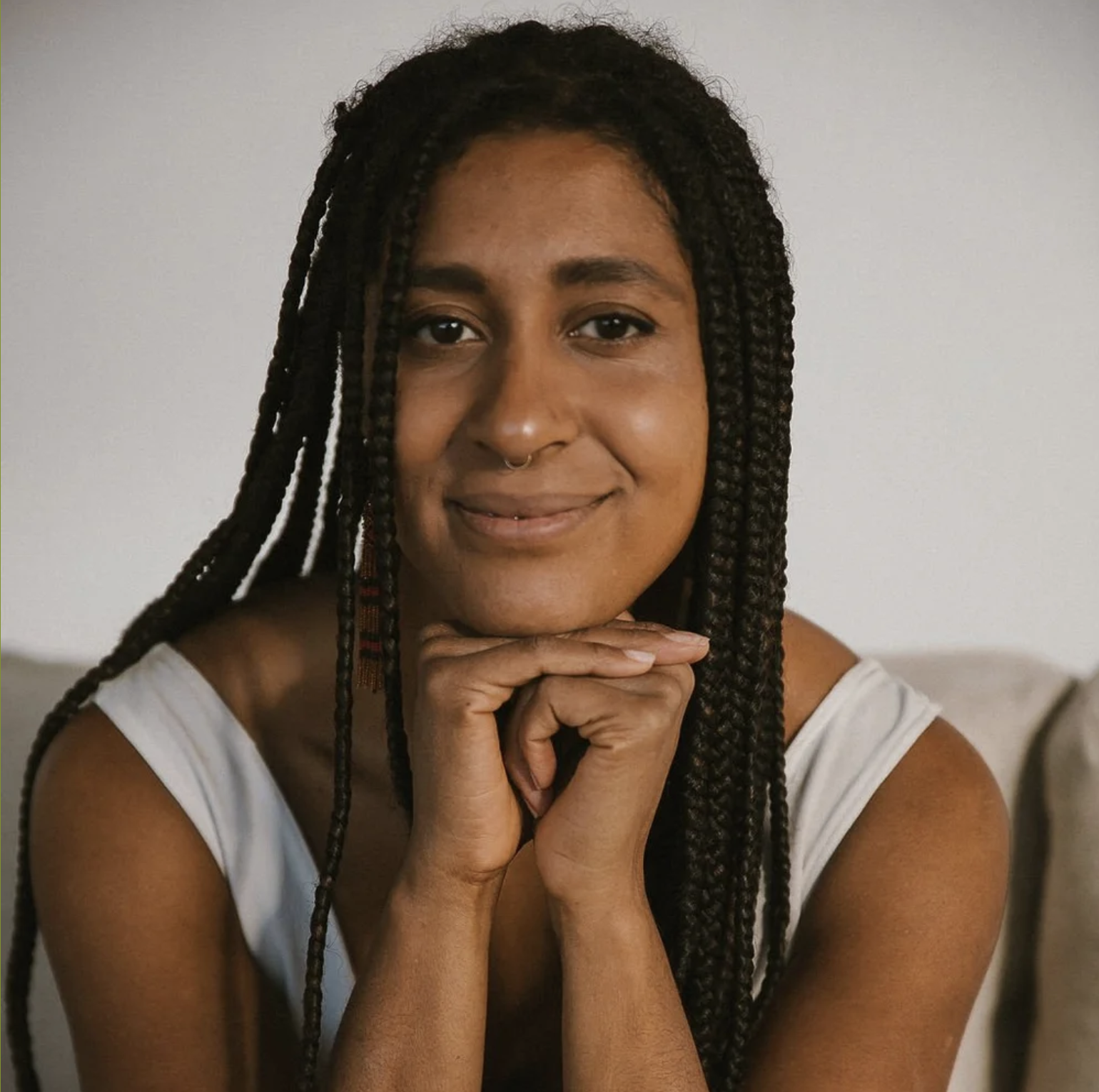 Portrait of a woman with braided hair, wearing a septum ring, smiling softly with her hands resting under her chin, wearing a sleeveless top, sitting against a neutral background.