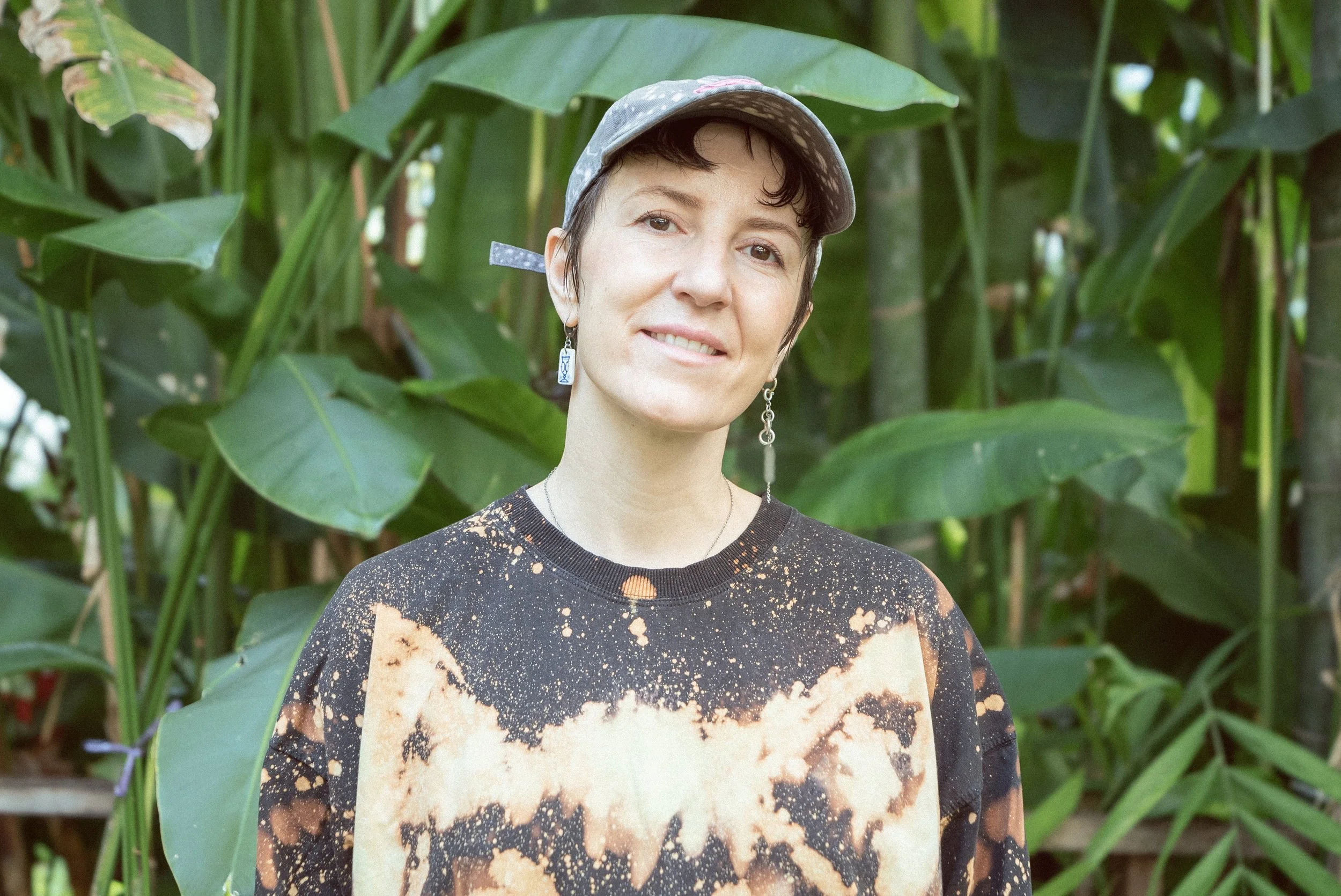 A person with short dark hair wearing a grey baseball cap, patterned earrings, and a tie-dye black and orange shirt, standing outdoors in front of green leafy plants.