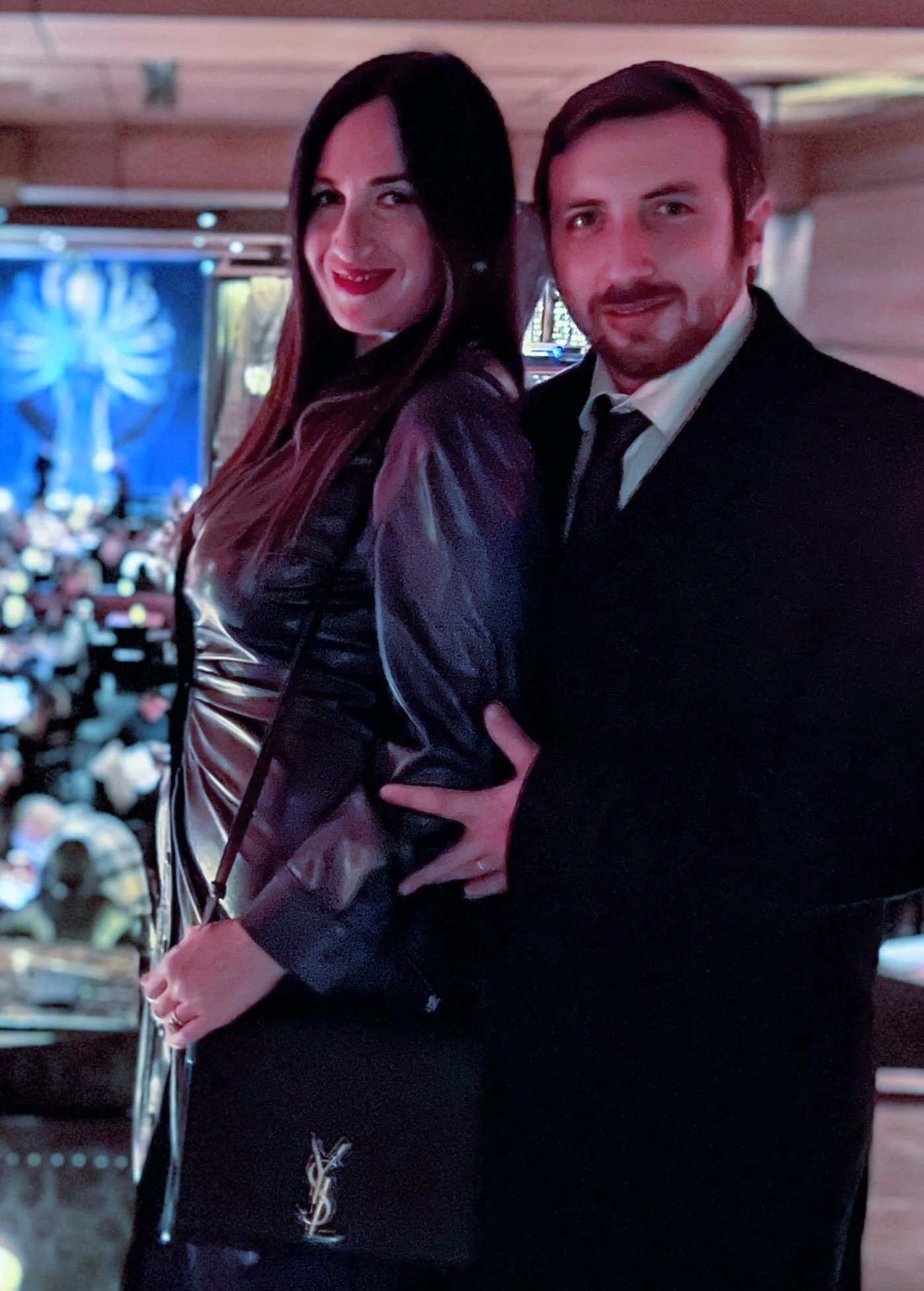 A man and woman dressed in formal attire, posing together indoors at an event, with a blurred audience and a large screen in the background.