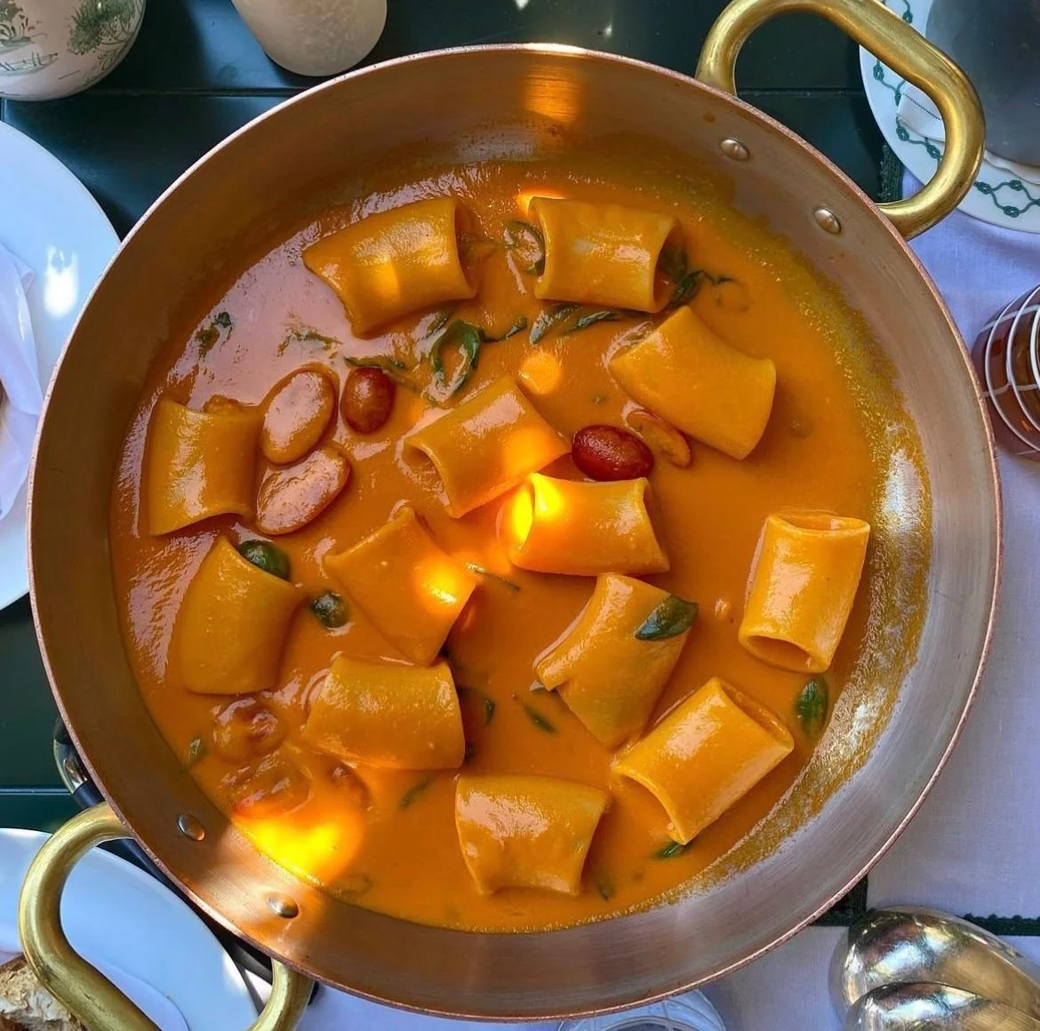 Pasta dish with large tubular noodles in a creamy orange sauce, topped with beans and herbs, in a large copper pan with gold handles.