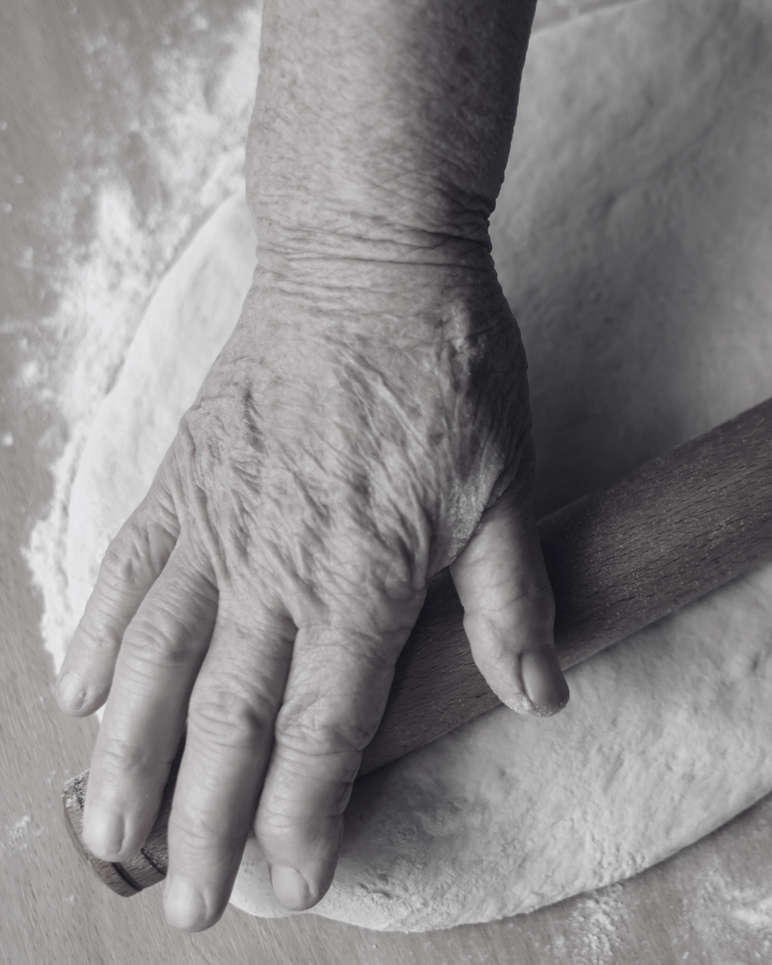 Close-up of an elderly person's hand rolling dough with a wooden rolling pin, dusted with flour.