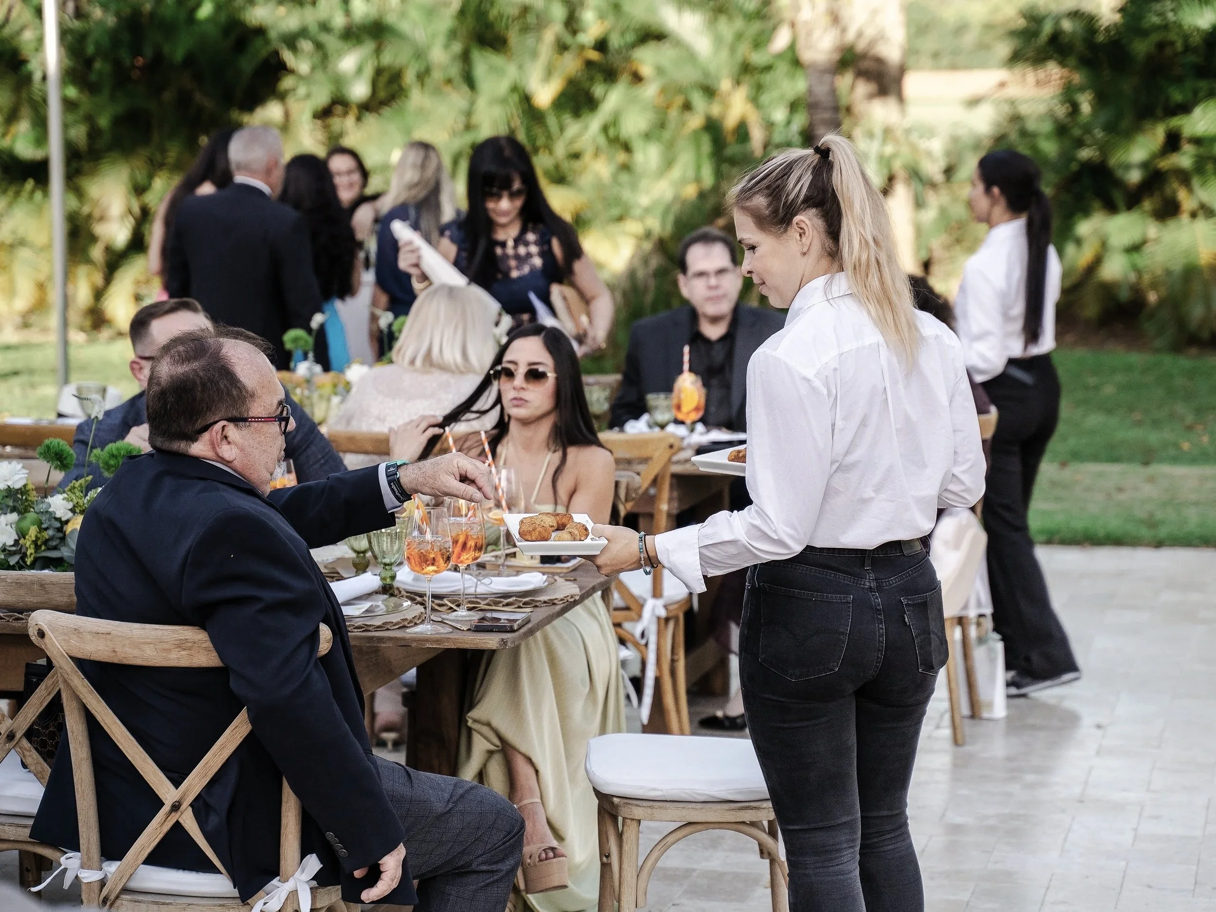 Servant girl serving food to a seated man at an outdoor gathering with other guests in the background.