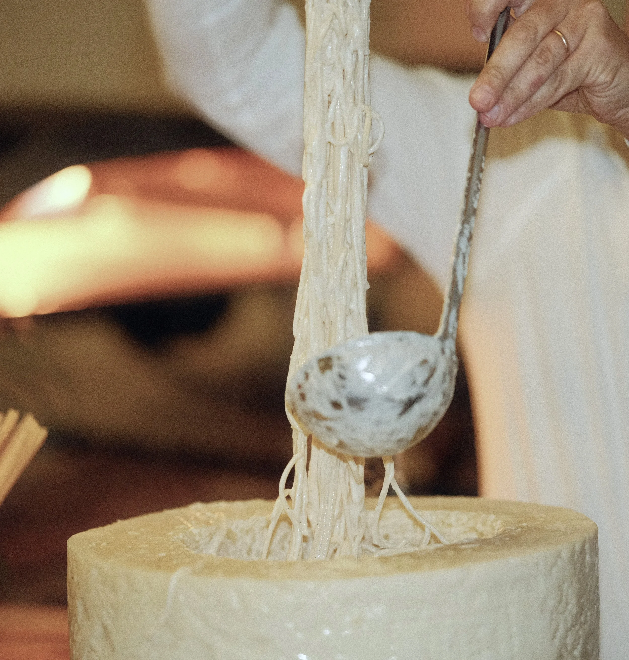 A person is scooping and stringing melted cheese from a wheel of cheese.