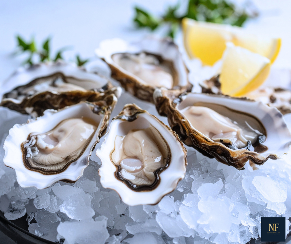 Six fresh oysters on the half shell served on ice with lemon wedges and green herbs in the background.