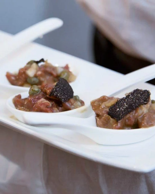 Three small servings of a savory dish with meat, vegetables, and a slice of black truffle on top, served on white ceramic spoons on a white rectangular plate.