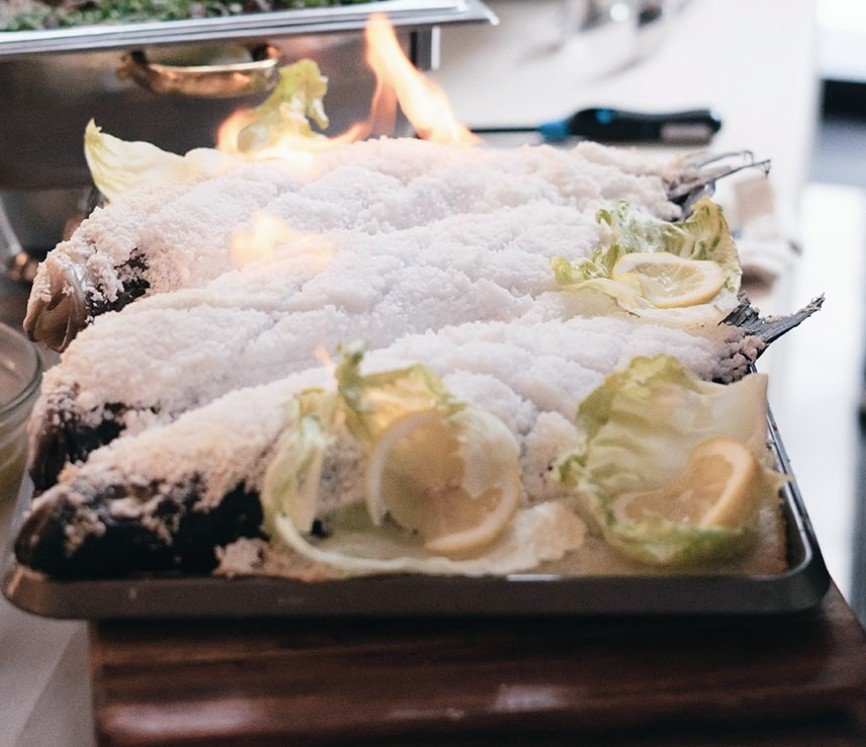 Whole fish coated with salt on a tray, garnished with lemon and lettuce leaves.