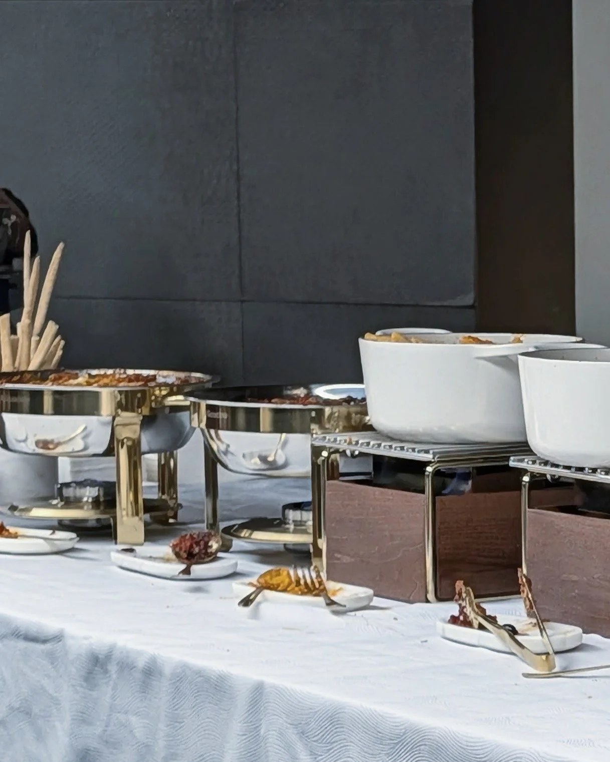Buffet table with dishes and utensils, including bowls and tongs on a white tablecloth against a dark wall.