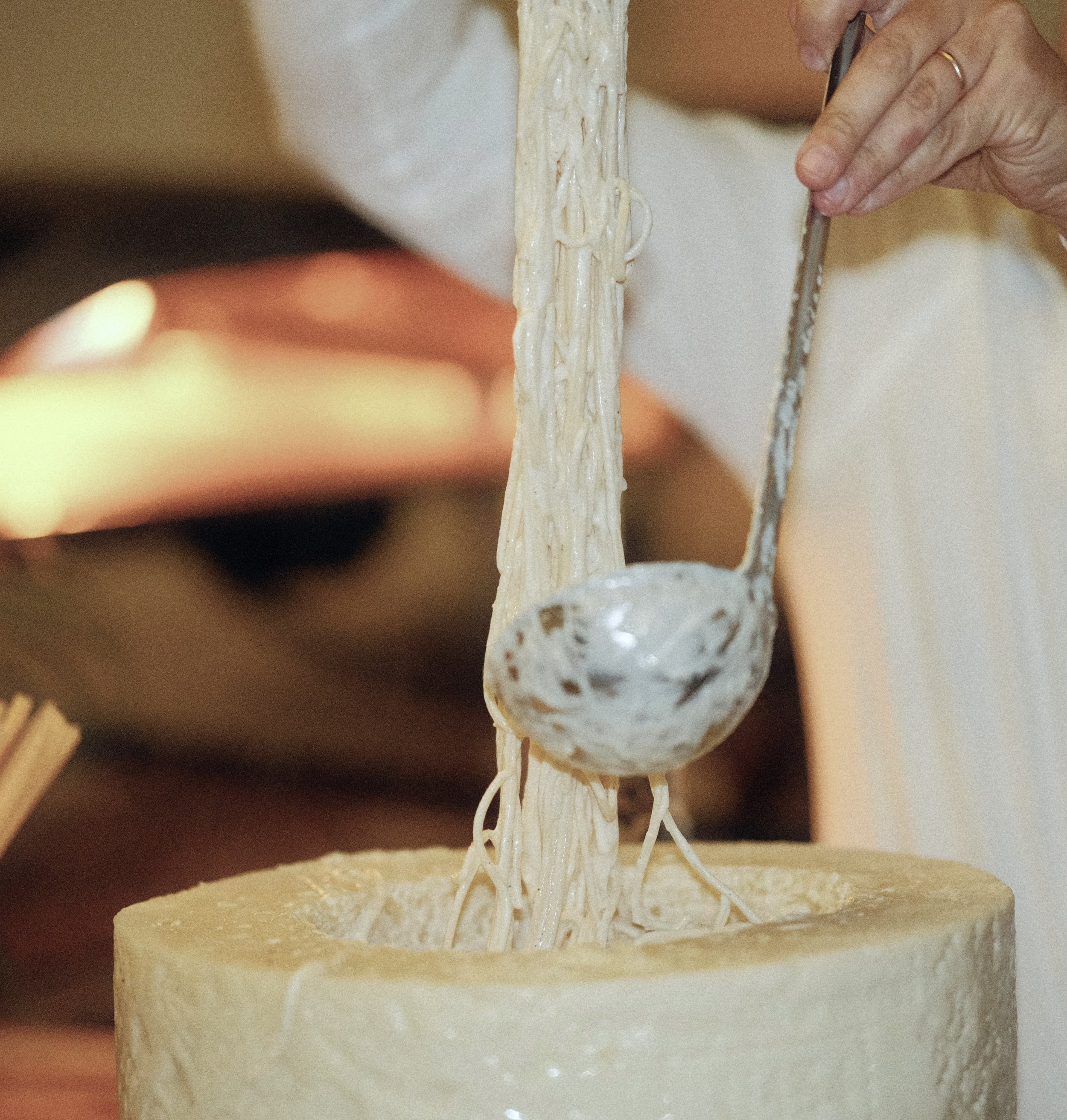 Close-up of a person using a spoon to lift and serve string cheese from a round cheese wheel.