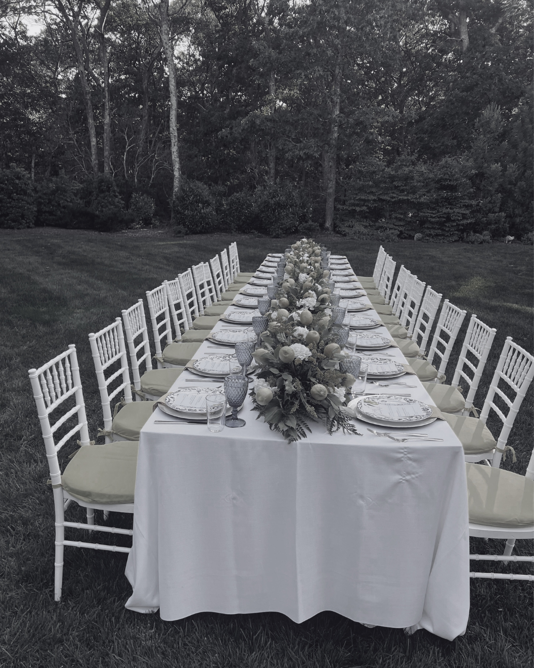 Dining table set for a formal outdoor event with white chairs, decorated with a lush floral centerpiece, plates, glasses, and silverware, arranged on a green lawn with trees in the background.