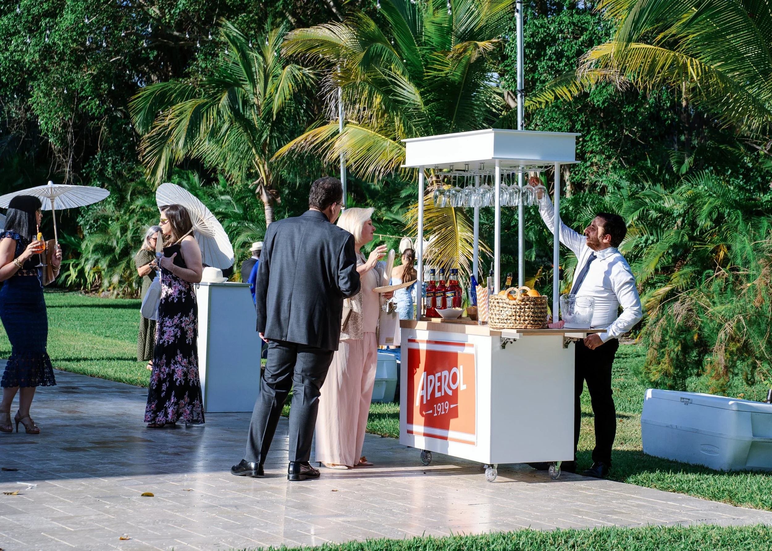People at an outdoor event enjoying drinks near a bar with Aperol branding, surrounded by green palm trees, with some holding umbrellas and sunglasses.