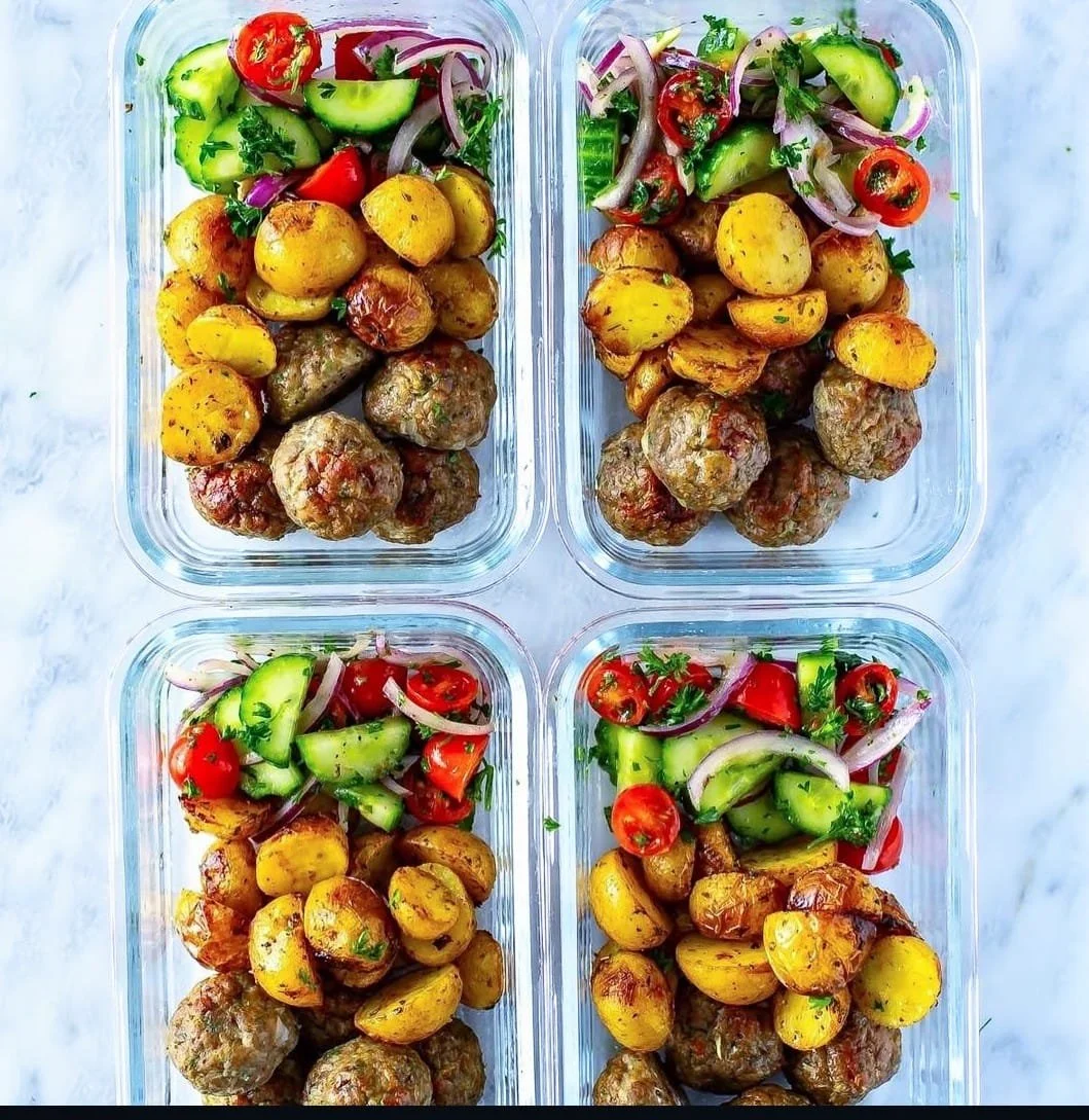 Four glass meal prep containers filled with roasted potatoes, meatballs, and vegetable salads including cucumbers, cherry tomatoes, red onions, and cilantro.