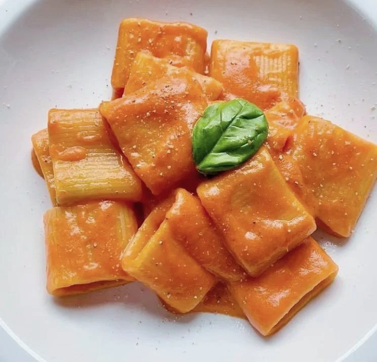 A white plate with cooked rigatoni pasta coated in orange marinara sauce, garnished with a single fresh basil leaf.