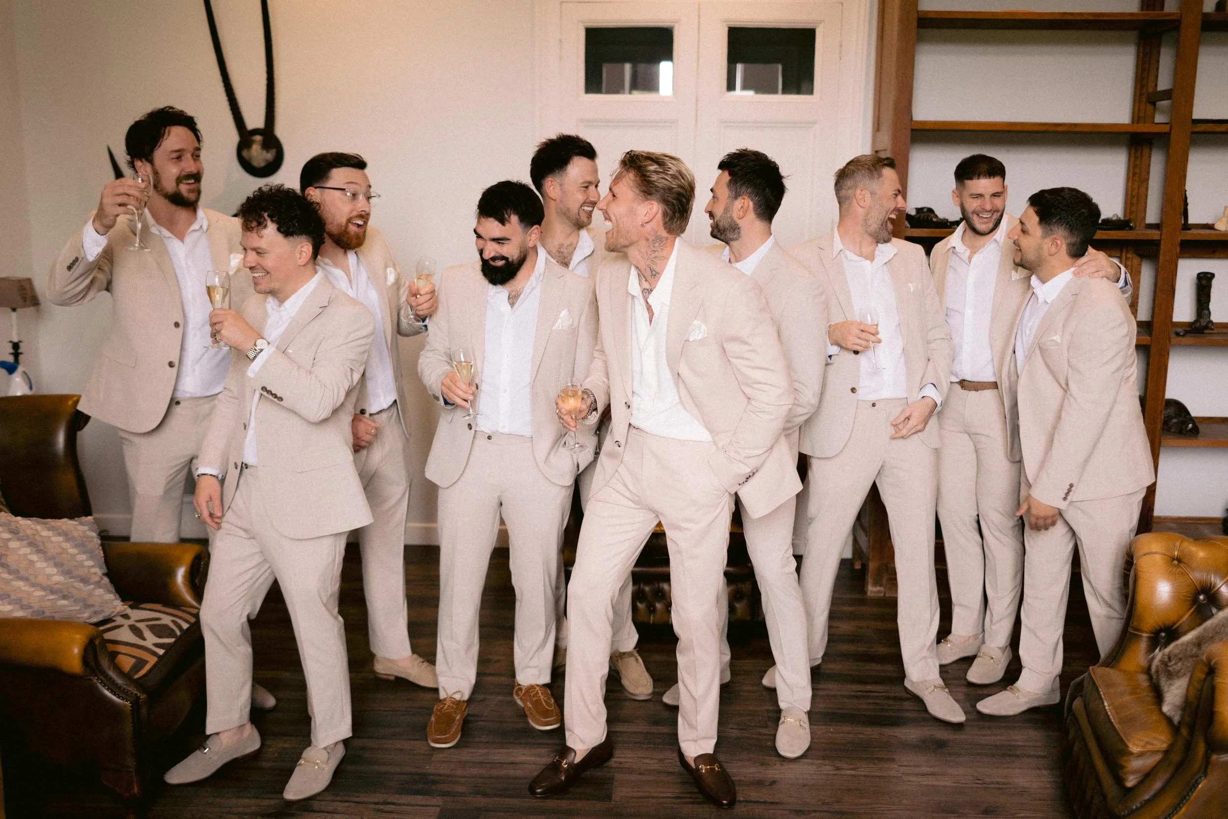 Group of men dressed in matching beige suits celebrating with drinks in a living room.