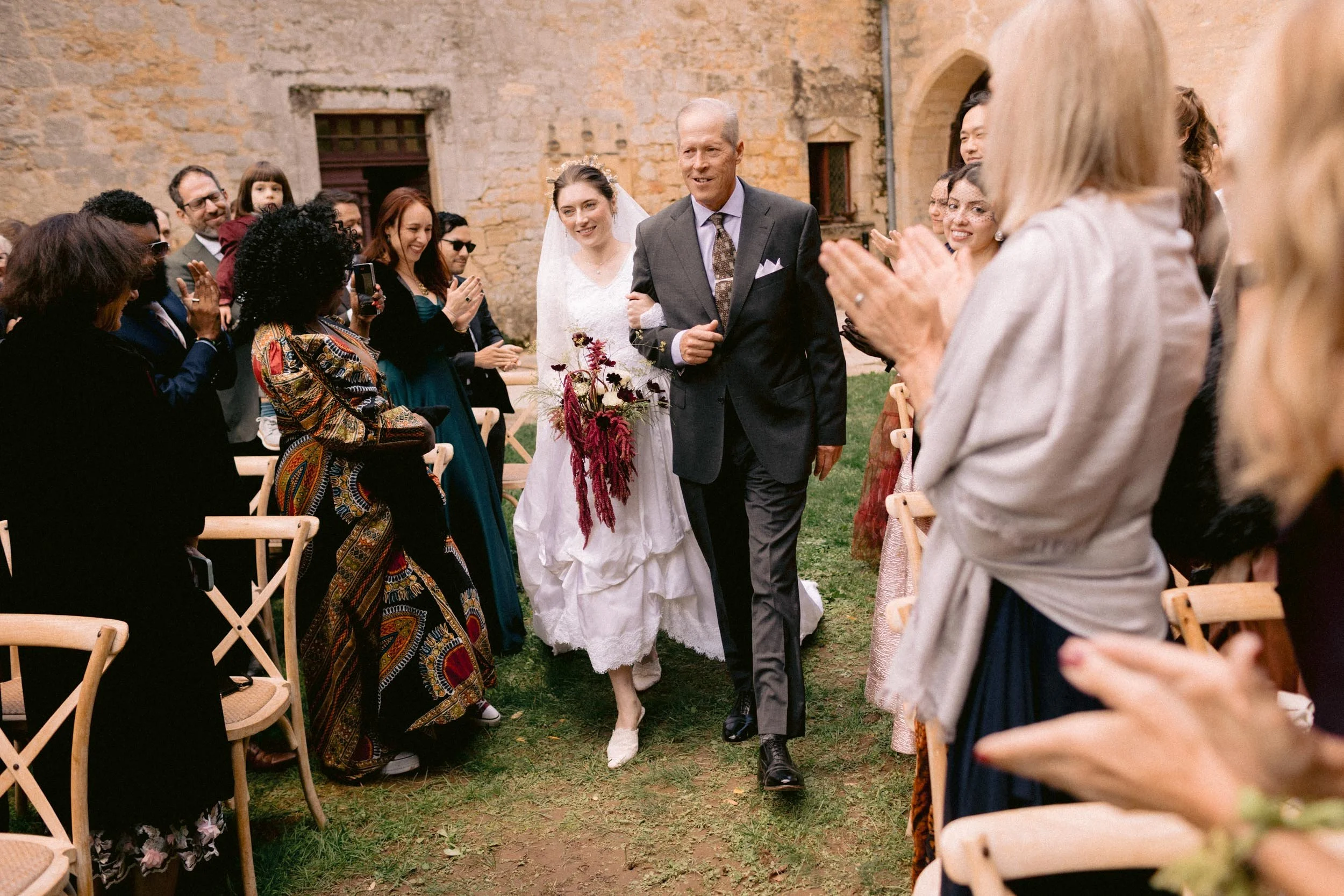A bride being walked down the aisle by her father at an outdoor wedding ceremony. Guests are clapping and smiling on both sides of the aisle, with a stone building in the background.