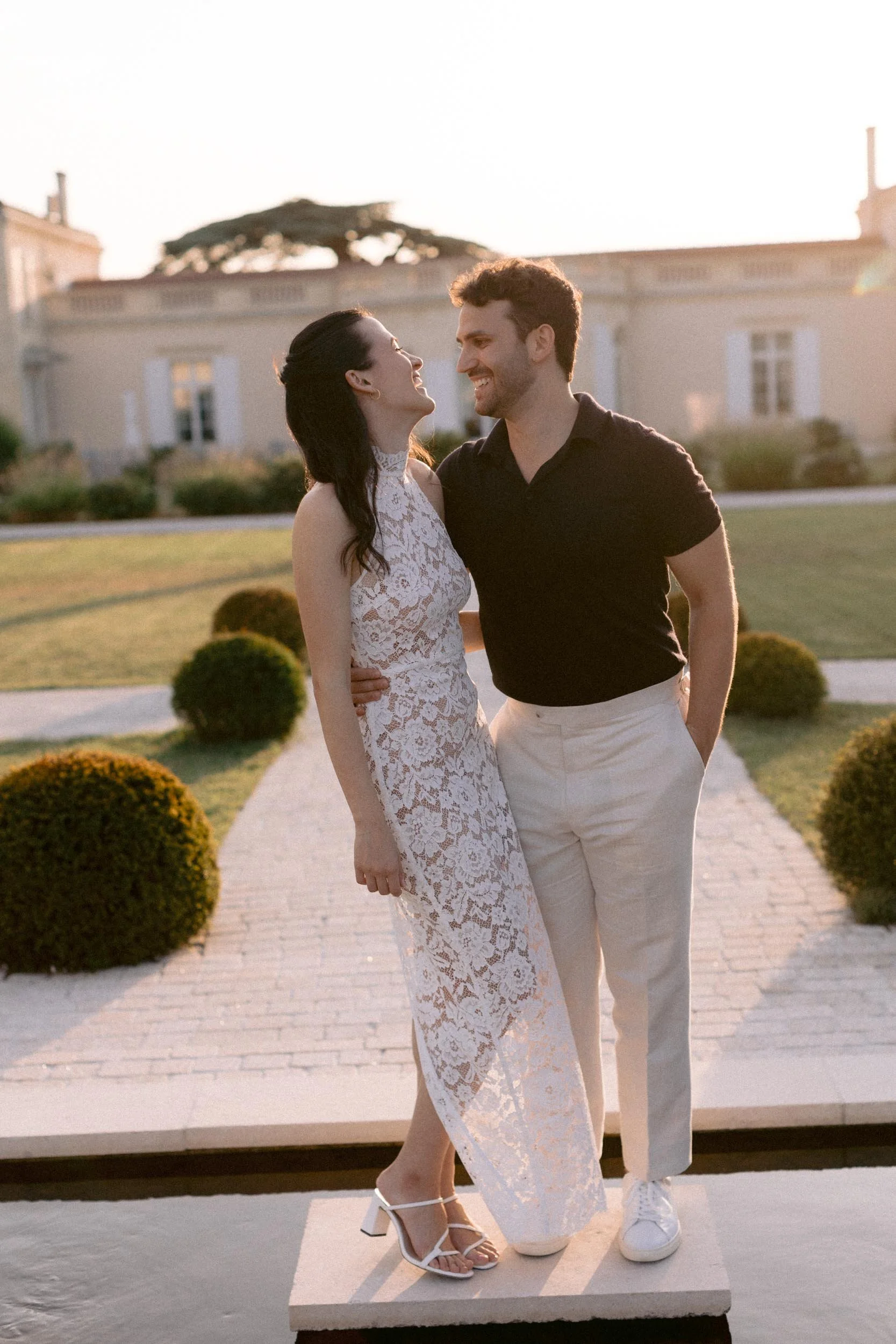 A couple smiling and looking at each other in an outdoor setting during sunset, with a large house and manicured bushes in the background.