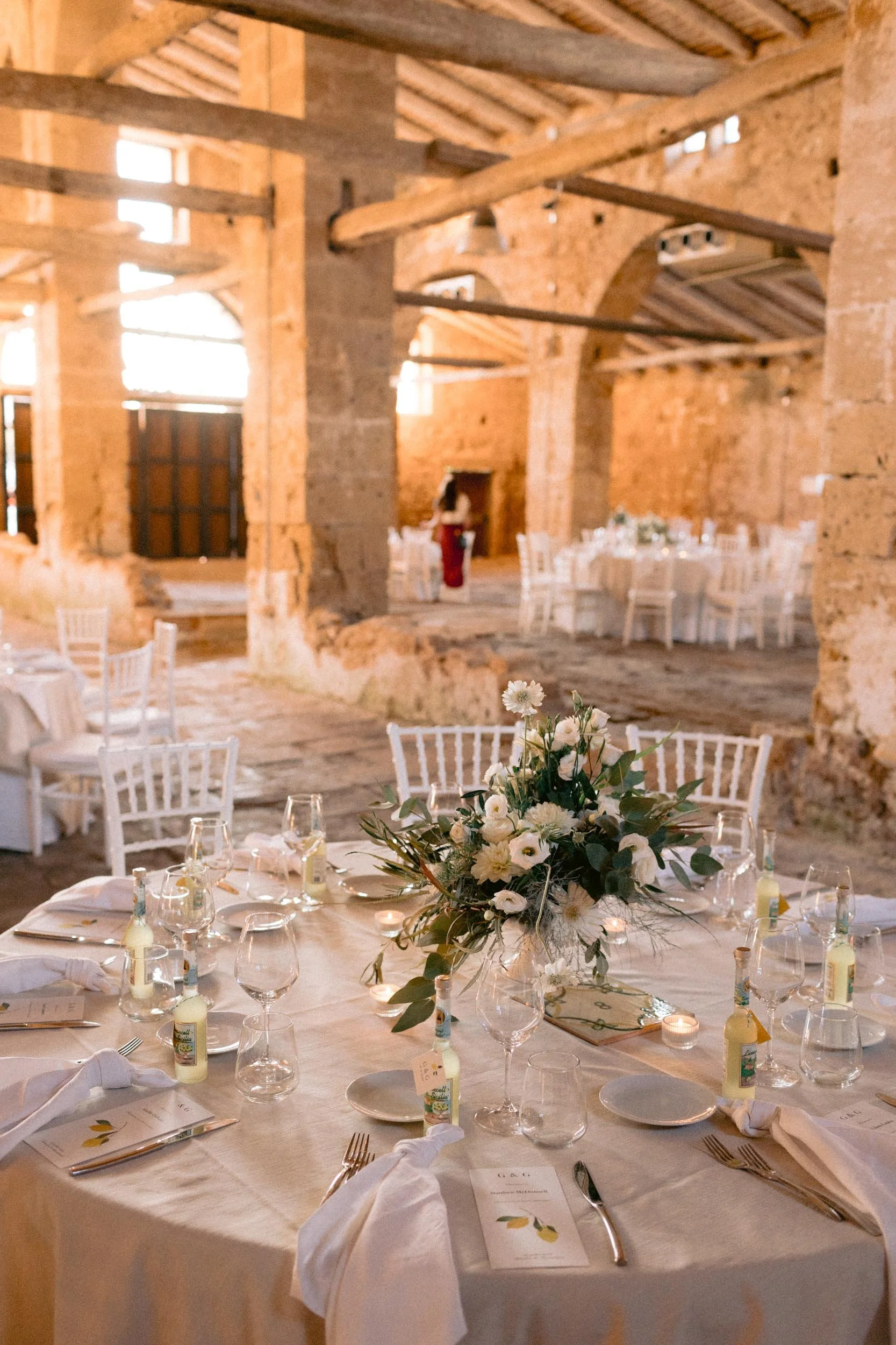 Inside a rustic wedding reception hall with stone walls and wooden beams, decorated with white chairs and tables, and a floral centerpiece on a round table set with plates, glasses, and white napkins.