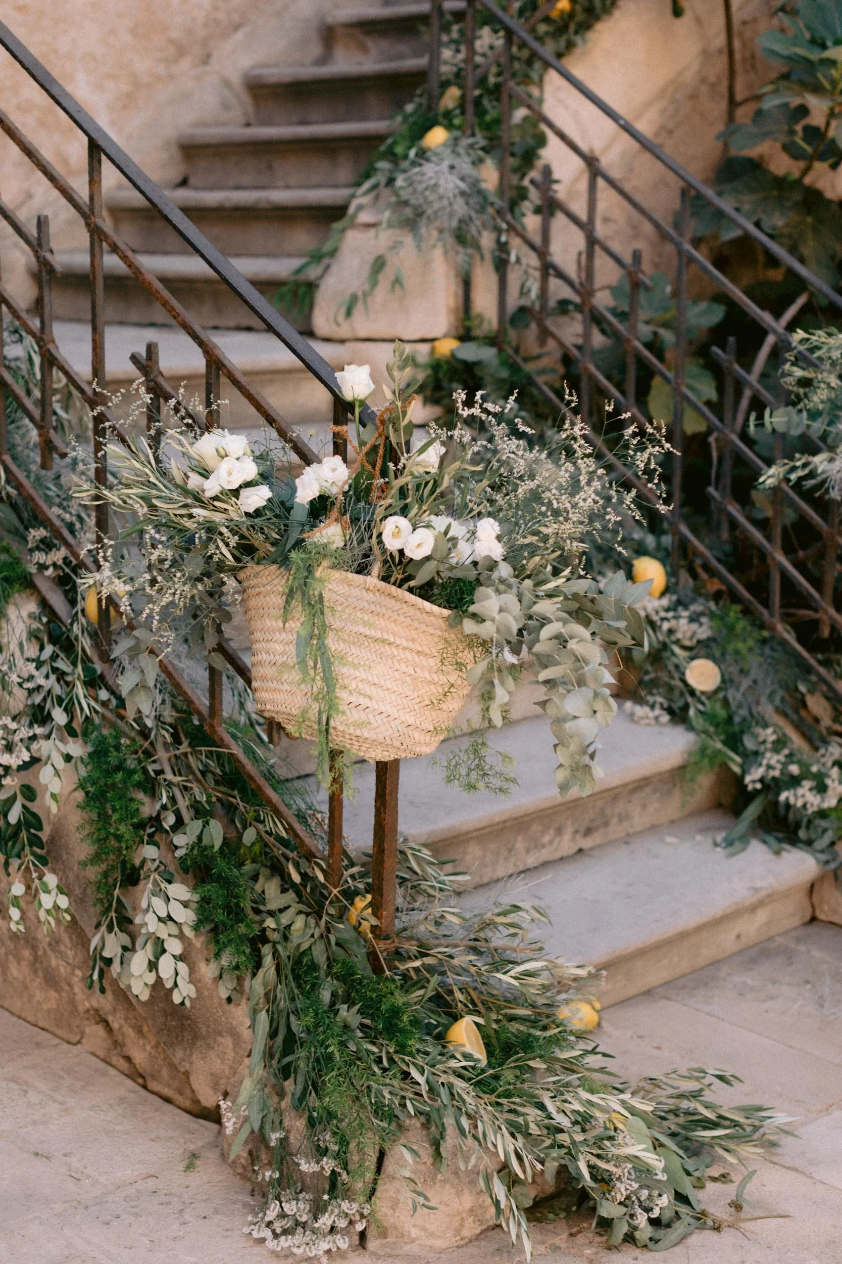 Decorated outdoor stone staircase with greenery, white flowers, and yellow accents, featuring a woven basket with floral arrangement hanging from the metal railing.