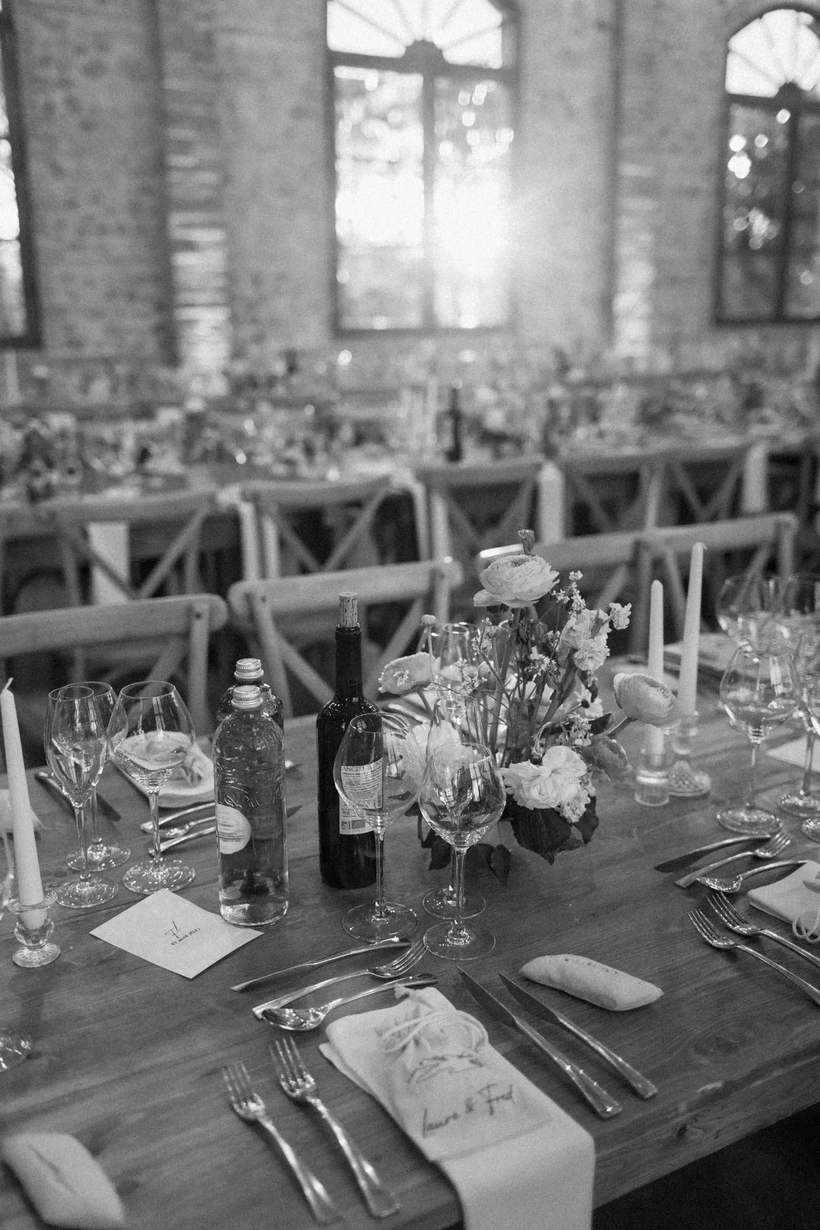 A black and white photo of a decorated dining table set for a formal event in a rustic brick-walled venue with large windows. The table features flowers, wine bottles, glasses, candles, and place settings with utensils and napkins.