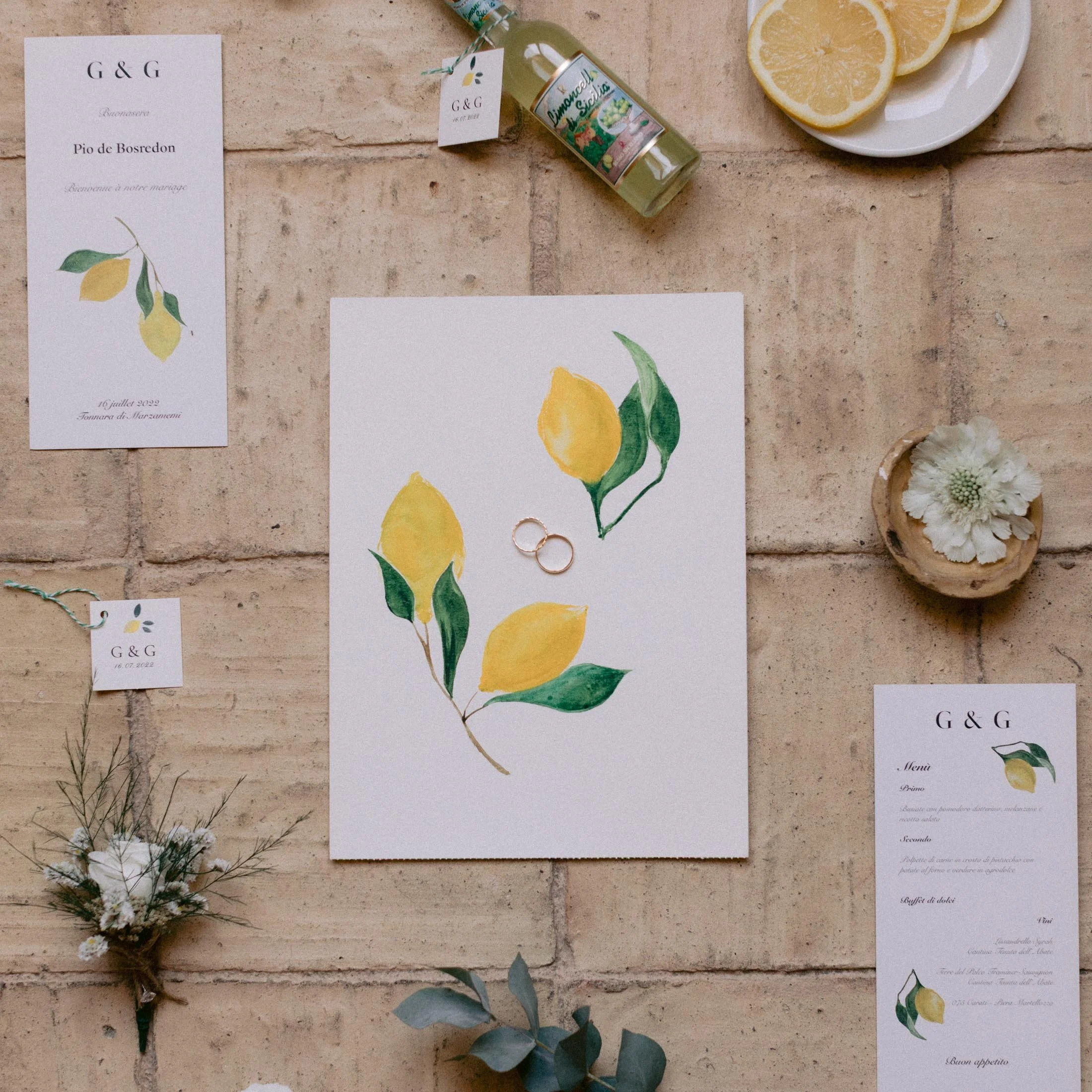Wedding invitation suite with illustrations of yellow lemons and green leaves, arranged on a brick surface along with small floral arrangements, a bottle, lemon slices, and gold wedding rings.