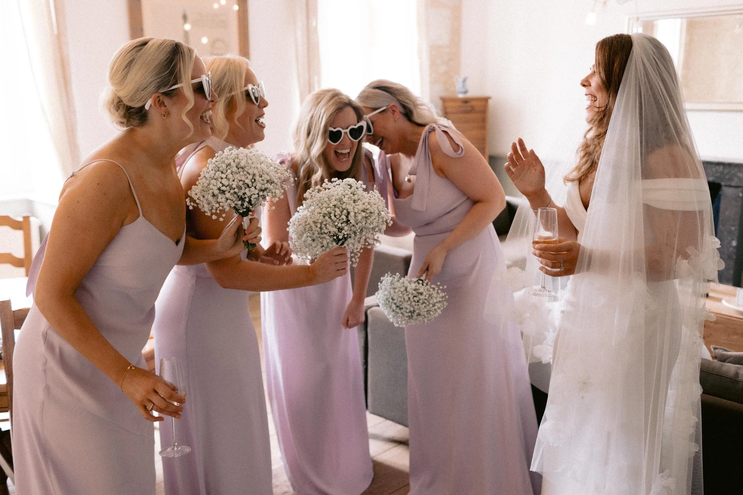 Bride in wedding dress holding a glass of champagne talking with five bridesmaids in lavender dresses, all smiling and holding white flowers, wearing sunglasses, in a warmly lit room.
