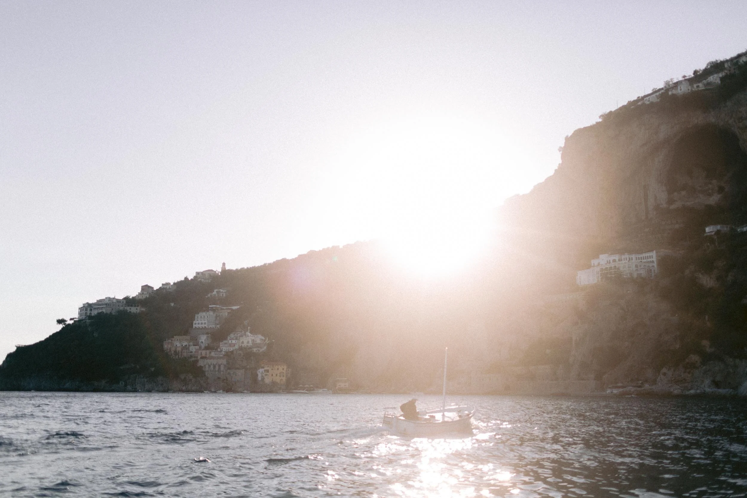 Sunset over a coastal hillside with houses, a boat on the water, and the sun shining brightly.