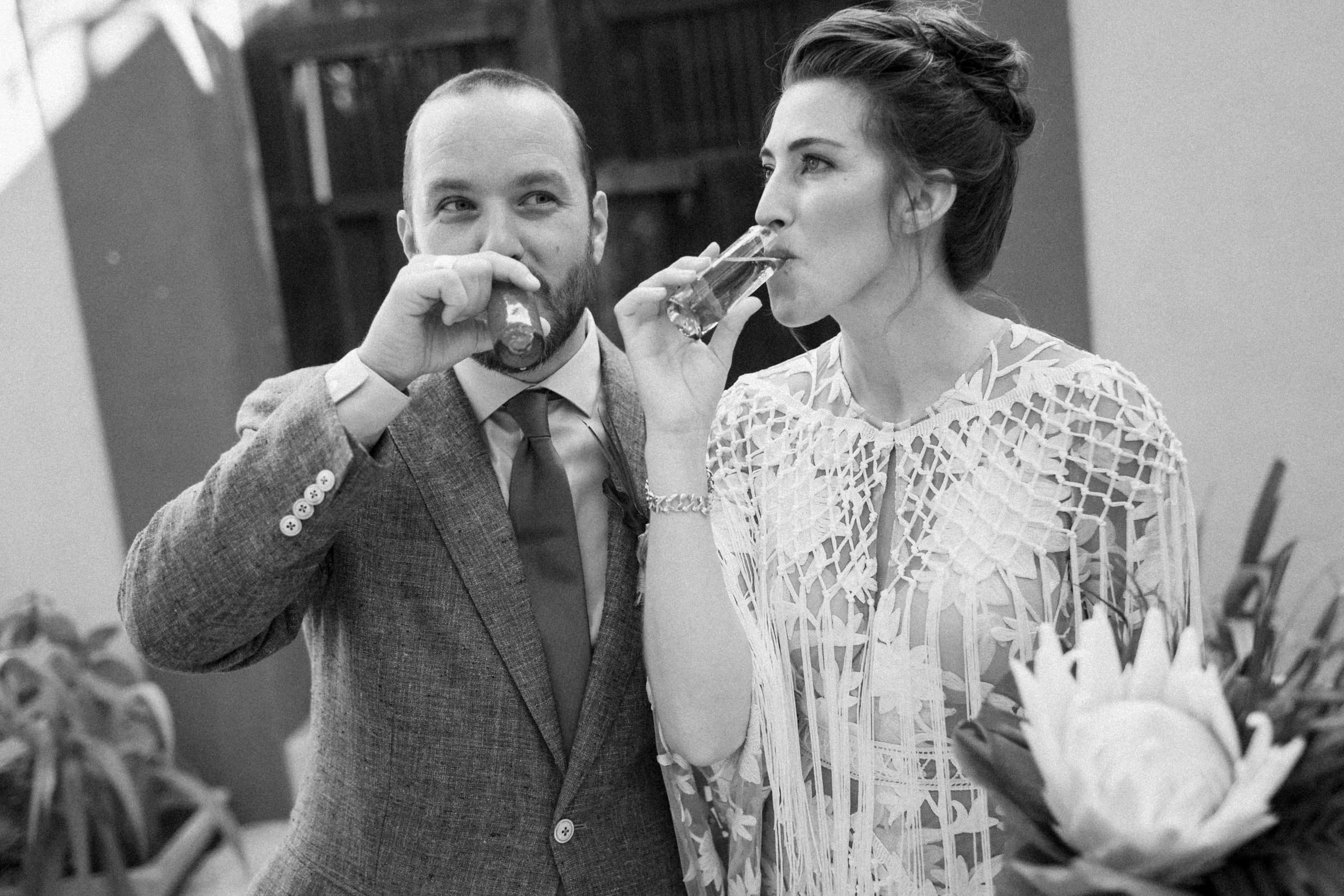 A man in a suit and a woman in a lace dress are drinking glasses of what appears to be champagne or wine. The man is looking at the woman while she looks forward, and they are standing near a table with flowers.