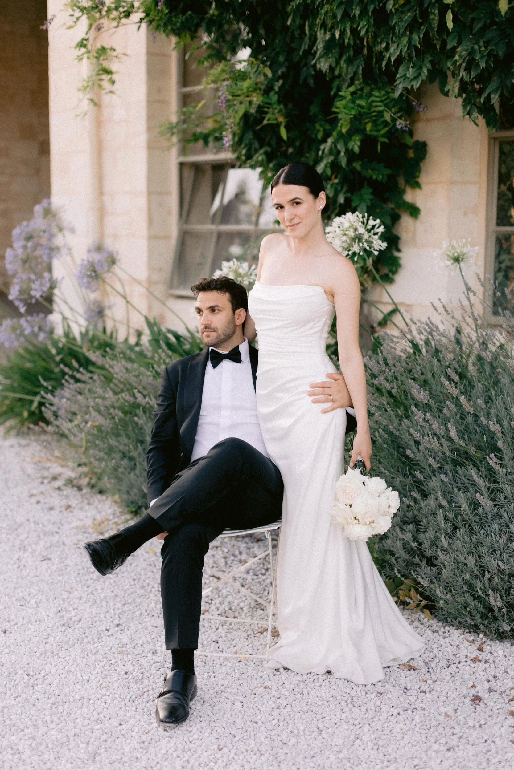 Bride in a white wedding dress holding a bouquet standing next to groom in a tuxedo sitting on a chair outdoors with greenery and flowers in the background.