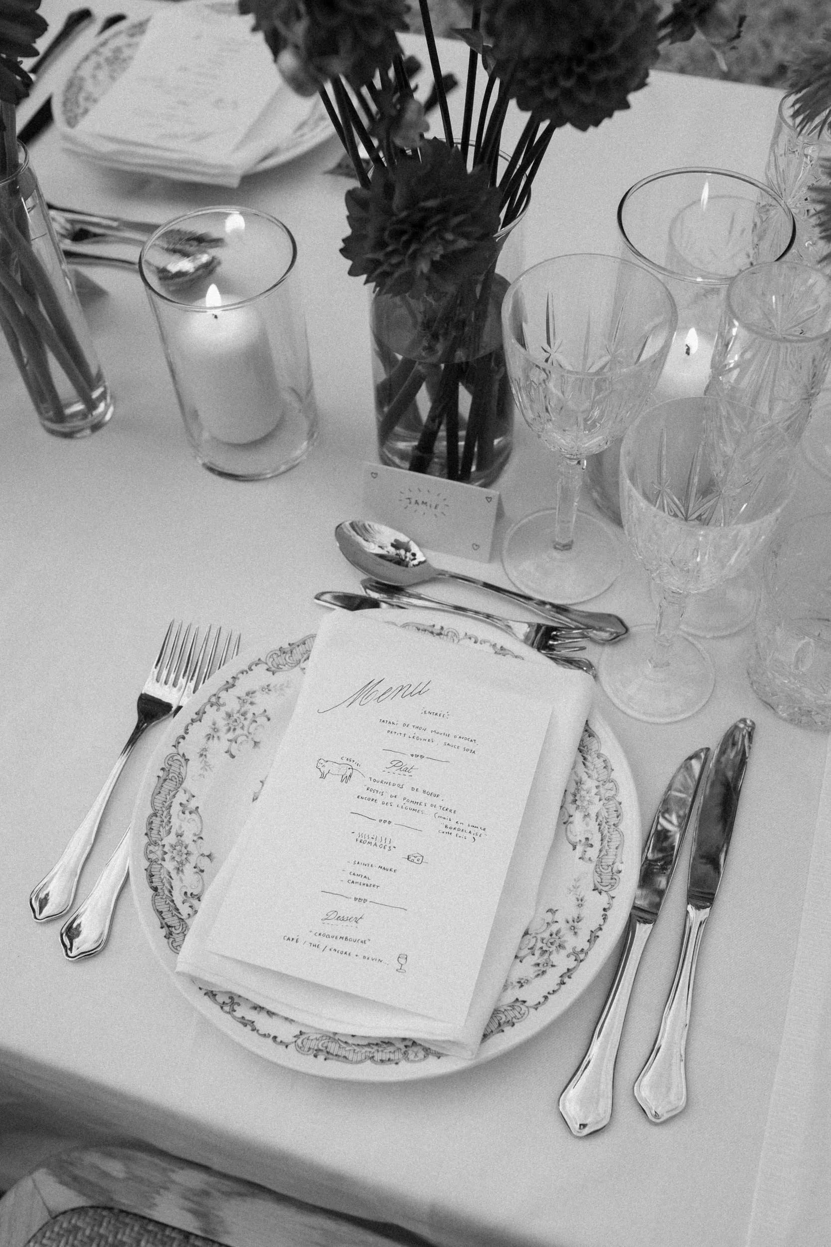 Set dining table with floral centerpiece, lit candles, crystal glasses, china plate, silverware, and a printed menu.