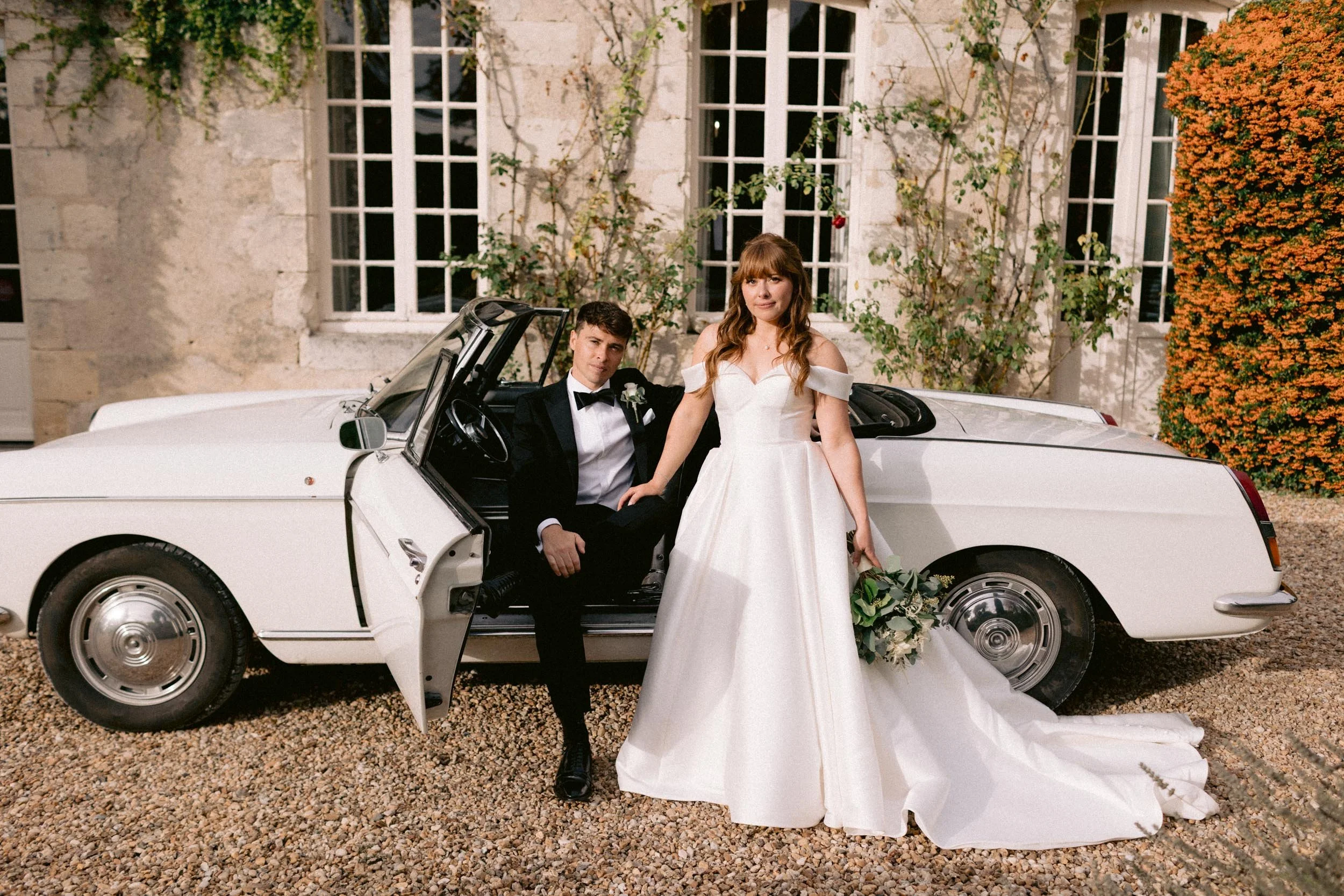 A bride and groom posing in front of a white vintage convertible car during their wedding. The bride is standing, holding a bouquet, while the groom is sitting inside the car, dressed in a tuxedo.