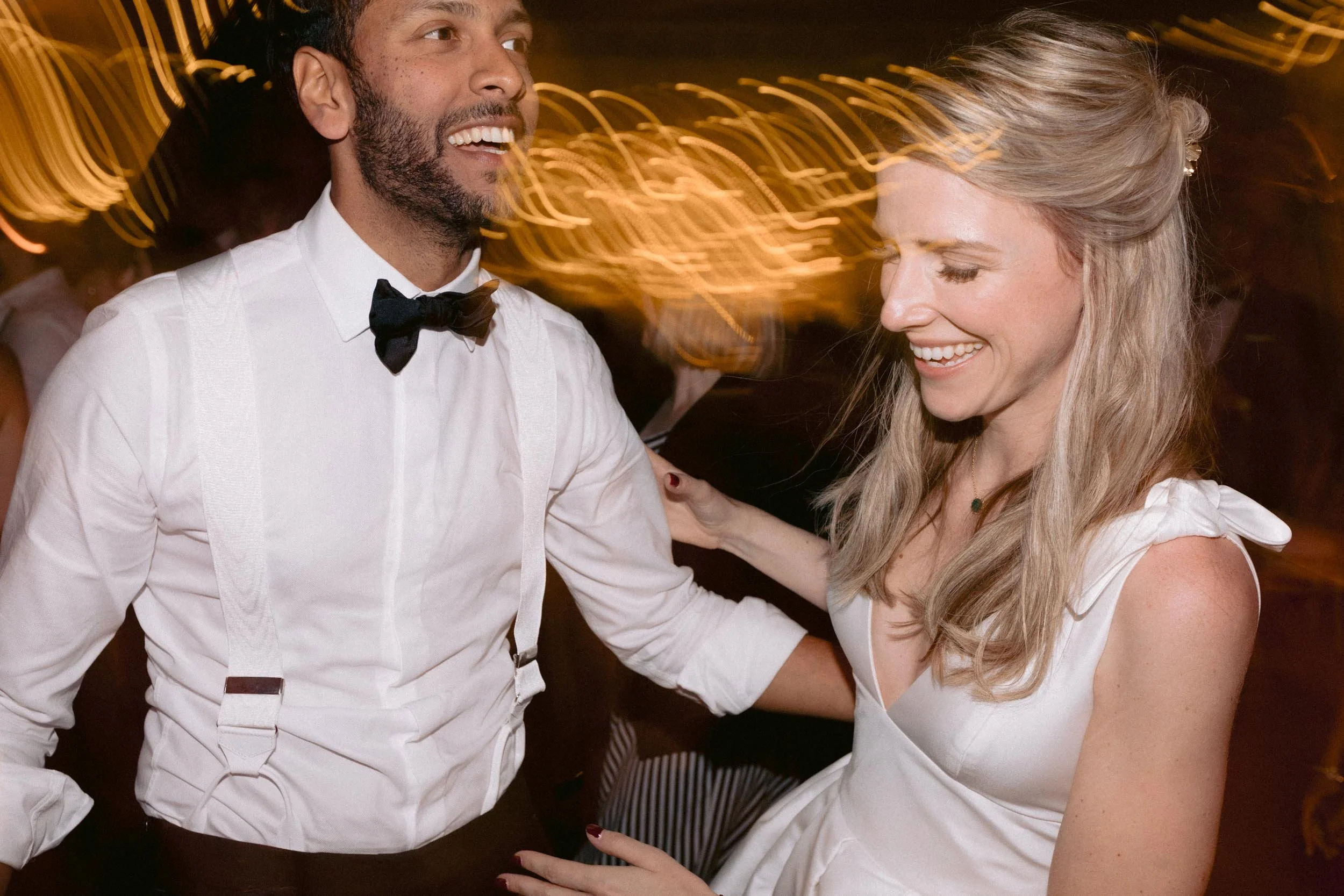 A bearded man in a white shirt with suspenders and a bow tie is dancing with a smiling woman in a white dress at a lively event, with warm, blurred lights in the background.