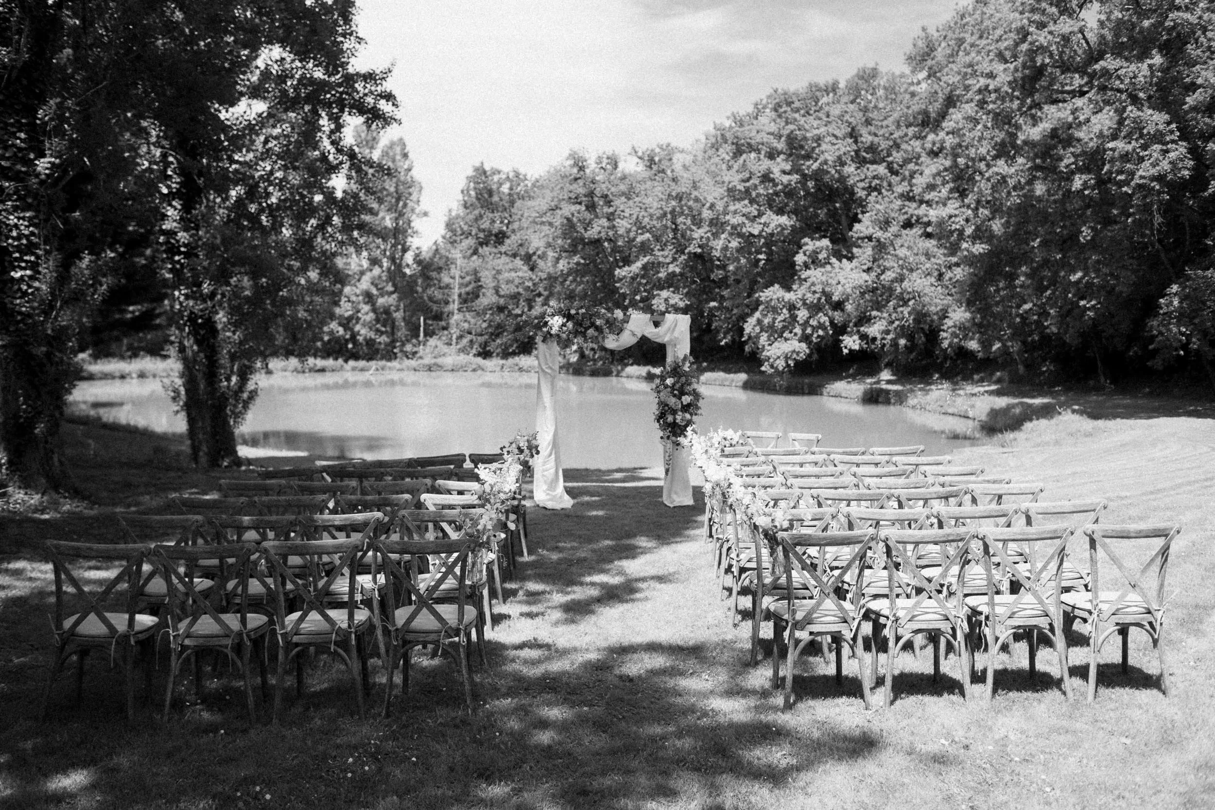 Outdoor wedding ceremony setup with chairs facing an arch decorated with flowers and fabric, situated by a lake with trees surrounding the area.