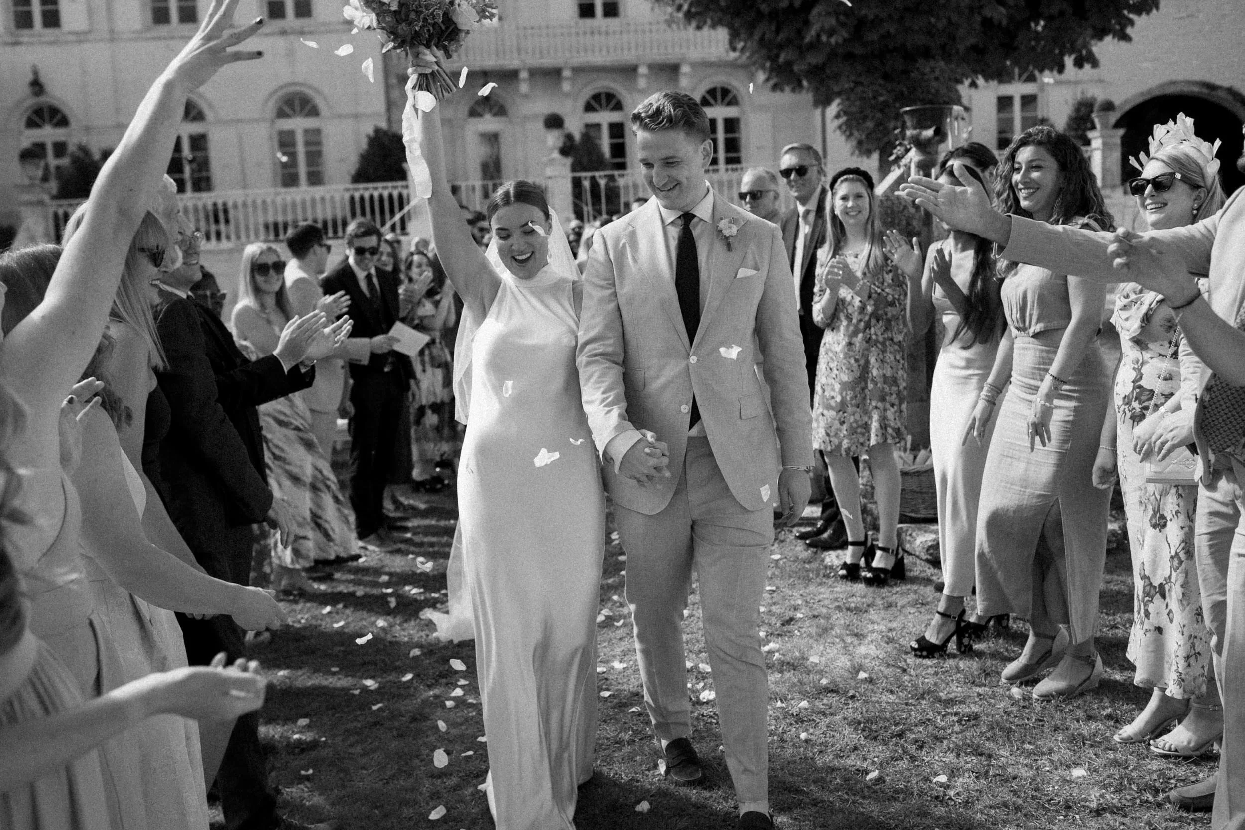 A bride and groom walking together outdoors, holding hands, surrounded by friends and family throwing petals during their wedding celebration.