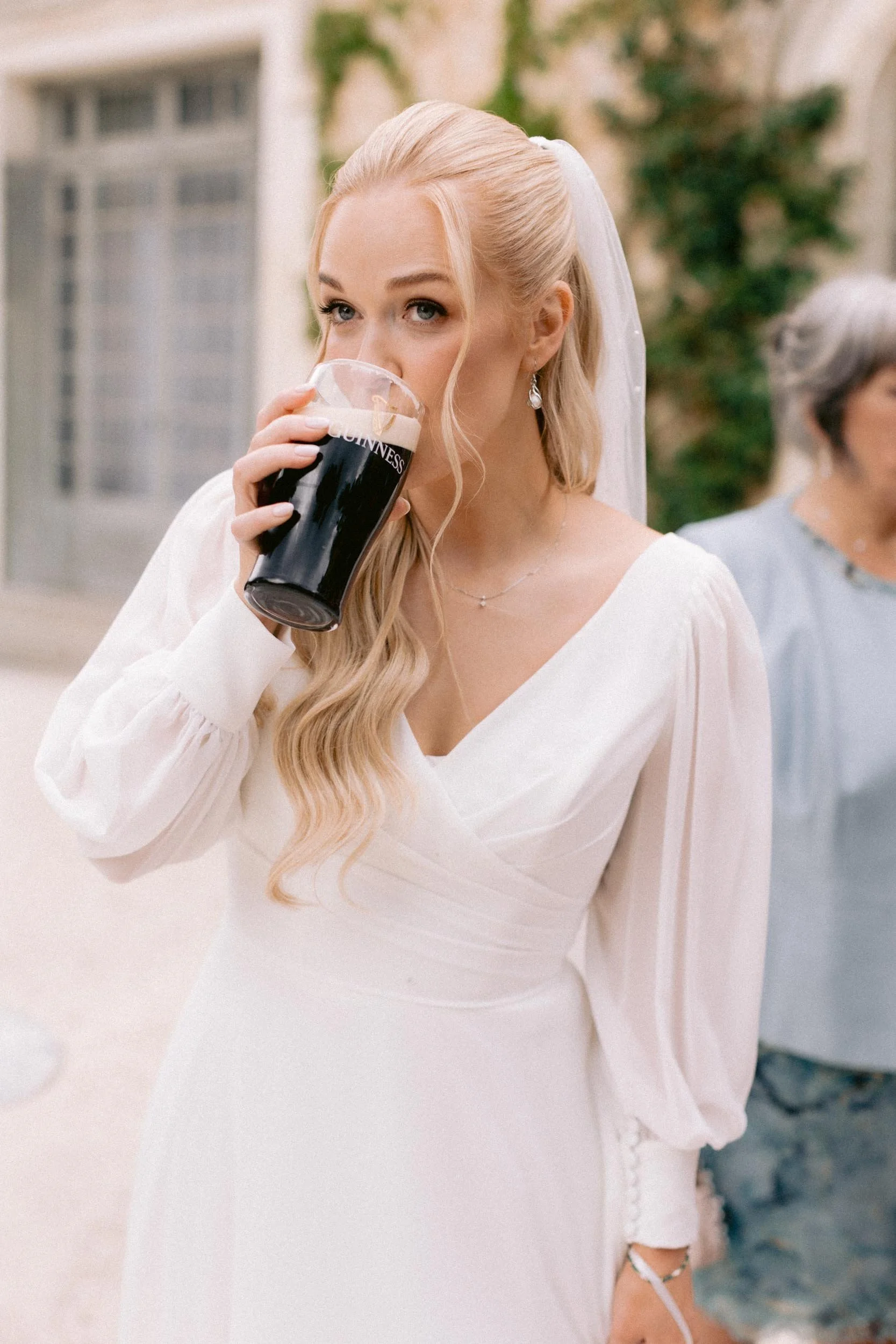 A bride in a white wedding dress drinking a pint of Guinness beer outdoors.