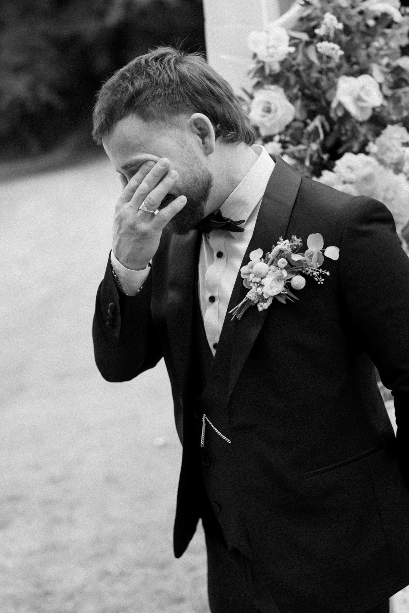 A groom in a tuxedo with a boutonniere, holding his face with his hand, appearing emotional at a wedding ceremony.