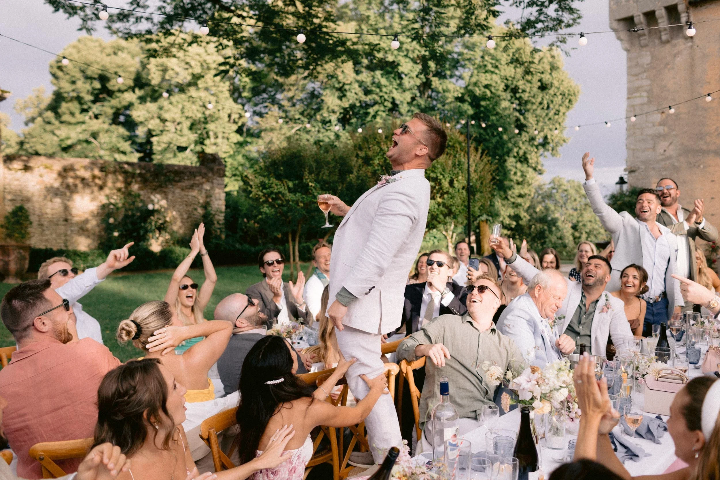 People celebrating outdoors at a wedding reception with trees and string lights in the background, some individuals are raising glasses, others cheering, and a man is standing on a chair with a glass of wine, laughing and enjoying the moment.