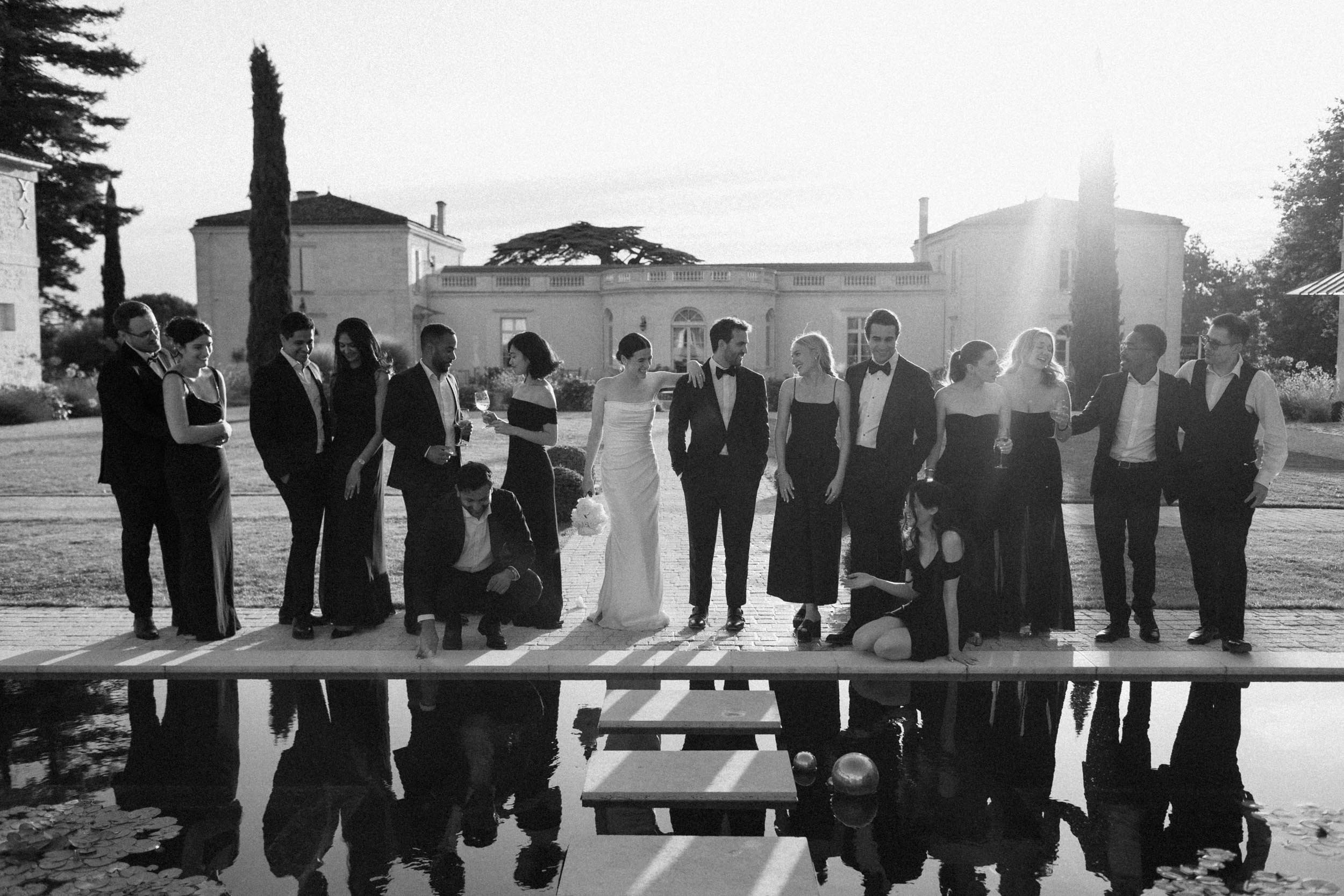 A black and white photo of a group of people dressed in formal attire standing by a pond with a bridge, outside a large estate or manor house during sunset.