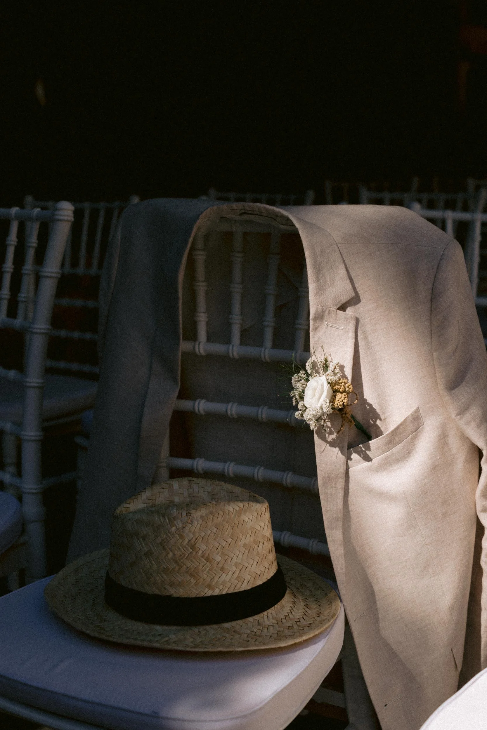 A beige suit jacket with a boutonniere hanging from the pocket, a straw hat with a black band resting on a white surface, and a black jacket draped over the back of a white chair at an elegant event.